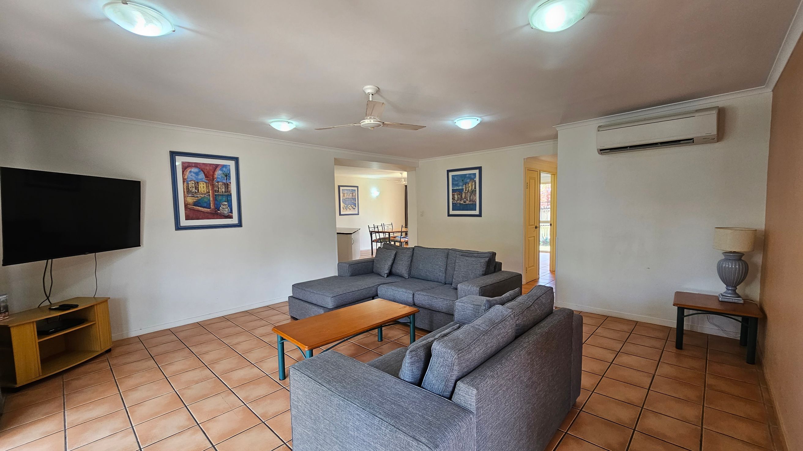 A living room with a tiled floor and a ceiling fan. On the left, a flat-screen TV is mounted on a wooden stand. In the centre, a grey L-shaped sofa faces a wooden coffee table. The right side features a side table with a lamp and an air conditioning unit above. The walls are adorned with framed artwork. In the background, a dining area with a table and chairs is visible through an open doorway. A living room with a tiled floor and a ceiling fan. On the left, a flat-screen TV is mounted on a wooden stand. In the centre, a grey L-shaped sofa faces a wooden coffee table. The right side features a side table with a lamp and an air conditioning unit above. The walls are adorned with framed artwork. In the background, a dining area with a table and chairs is visible through an open doorway.