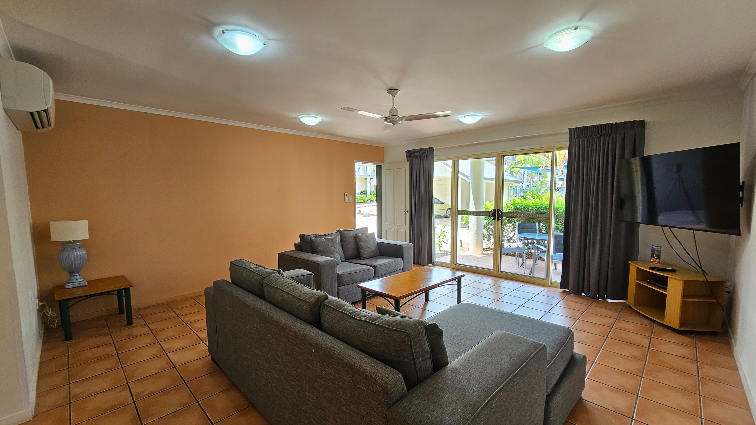 A living room with terracotta tiled flooring and a white ceiling featuring recessed lights and a ceiling fan. On the left, there's a small table with a lamp and an air conditioning unit above. In the centre, two grey sofas face a wooden coffee table. The right side has a wall-mounted TV on a wooden stand. Large glass doors with dark curtains lead to an outdoor area with visible greenery and a table with chairs. A living room with terracotta tiled flooring and a white ceiling featuring recessed lights and a ceiling fan. On the left, there's a small table with a lamp and an air conditioning unit above. In the centre, two grey sofas face a wooden coffee table. The right side has a wall-mounted TV on a wooden stand. Large glass doors with dark curtains lead to an outdoor area with visible greenery and a table with chairs.