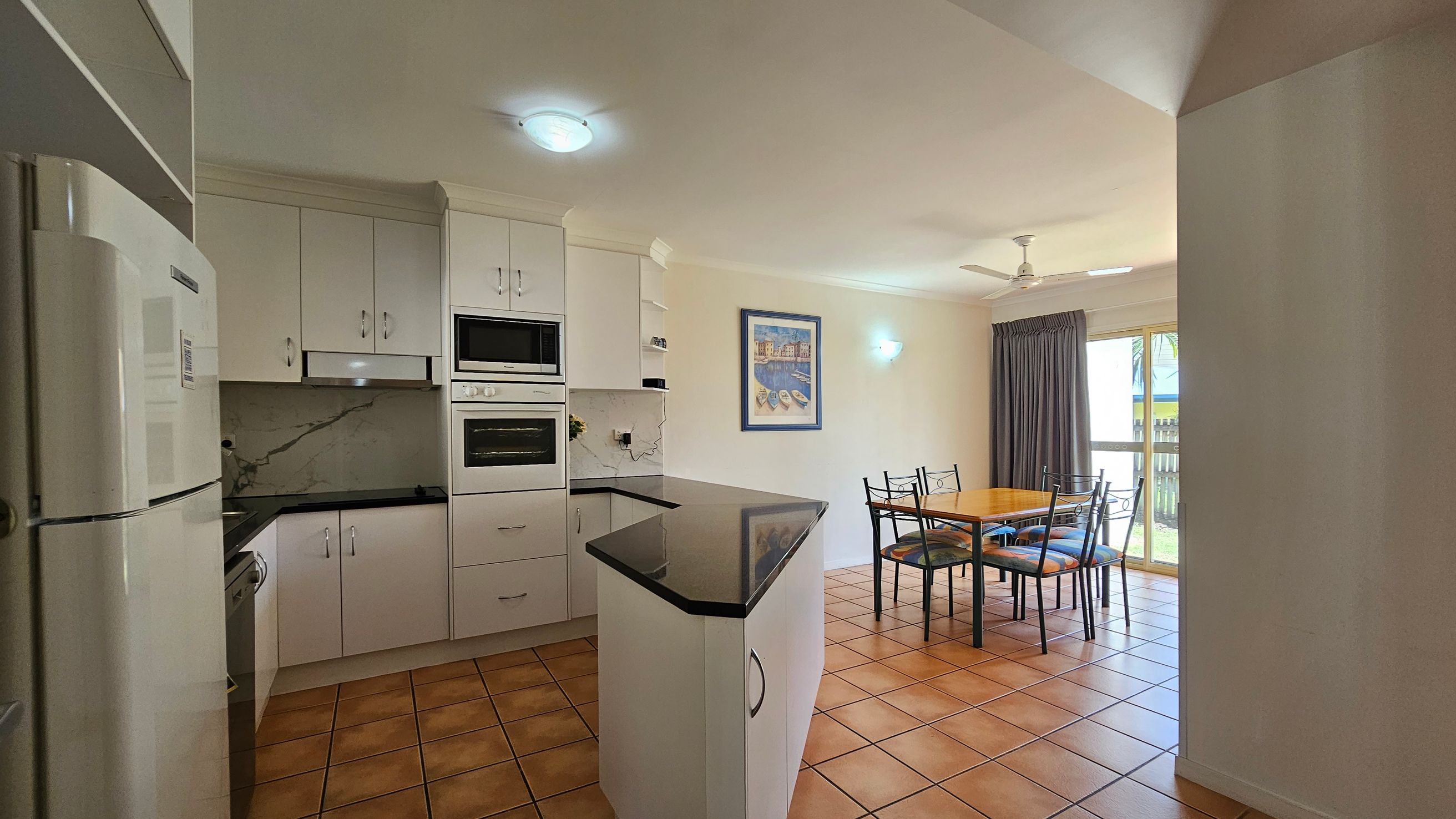 A kitchen and dining area with terracotta floor tiles. On the left, there is a white refrigerator and cabinetry with a built-in oven and microwave. A black countertop extends into the room. In the middle, a dining table with four chairs is set against a wall with a framed picture. On the right, a large window with dark curtains and a ceiling fan above. The ceiling is white with recessed lighting. A kitchen and dining area with terracotta floor tiles. On the left, there is a white refrigerator and cabinetry with a built-in oven and microwave. A black countertop extends into the room. In the middle, a dining table with four chairs is set against a wall with a framed picture. On the right, a large window with dark curtains and a ceiling fan above. The ceiling is white with recessed lighting.
