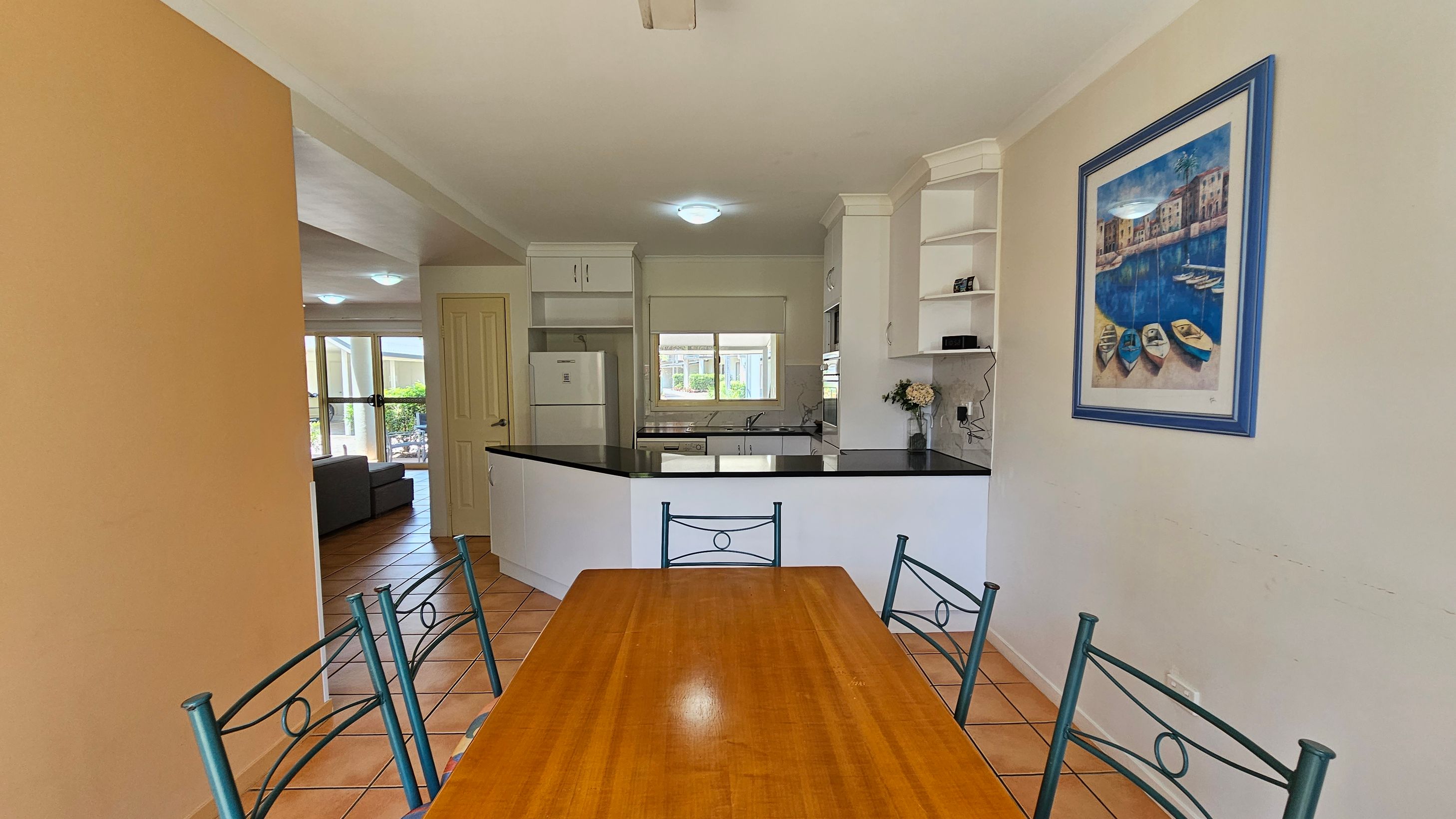 A dining area with a wooden table and metal chairs in the foreground. To the right, a framed painting of boats on water hangs on the wall. In the background, a kitchen with white cabinets, a black countertop, and a window. A refrigerator is visible to the left of the kitchen. Beyond the kitchen, a living area with a grey sofa and large windows can be seen. The floor is tiled with a terracotta colour. A dining area with a wooden table and metal chairs in the foreground. To the right, a framed painting of boats on water hangs on the wall. In the background, a kitchen with white cabinets, a black countertop, and a window. A refrigerator is visible to the left of the kitchen. Beyond the kitchen, a living area with a grey sofa and large windows can be seen. The floor is tiled with a terracotta colour.