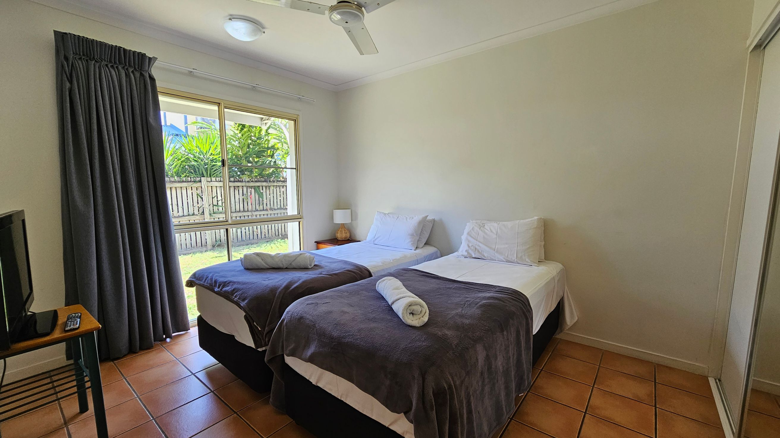 A bedroom with two single beds in the centre, each with white sheets, pillows, and a rolled towel on top of dark blankets. A small bedside table with a lamp is between the beds. On the left, a window with closed dark curtains overlooks a garden with a wooden fence and greenery. A TV sits on a small table with a remote control. The floor is tiled in a terracotta colour, and a ceiling fan is visible above. A mirrored wardrobe is on the right. A bedroom with two single beds in the centre, each with white sheets, pillows, and a rolled towel on top of dark blankets. A small bedside table with a lamp is between the beds. On the left, a window with closed dark curtains overlooks a garden with a wooden fence and greenery. A TV sits on a small table with a remote control. The floor is tiled in a terracotta colour, and a ceiling fan is visible above. A mirrored wardrobe is on the right.