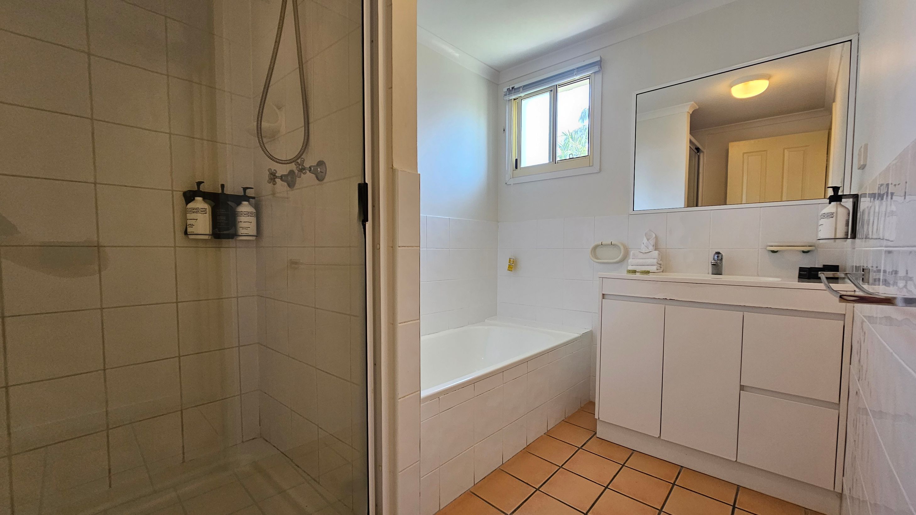 A bathroom with a tiled floor and white walls. On the left, there's a glass-enclosed shower with mounted soap dispensers. In the centre, a white bathtub is positioned beneath a window with a blind. On the right, a white vanity unit with a large mirror above it holds a soap dispenser and a small decorative item. The ceiling light is visible in the mirror's reflection. A bathroom with a tiled floor and white walls. On the left, there's a glass-enclosed shower with mounted soap dispensers. In the centre, a white bathtub is positioned beneath a window with a blind. On the right, a white vanity unit with a large mirror above it holds a soap dispenser and a small decorative item. The ceiling light is visible in the mirror's reflection.
