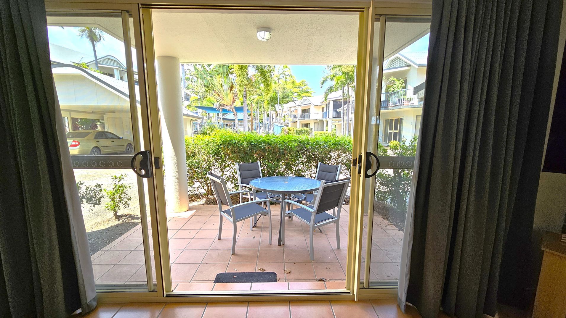 Sliding glass doors open to a patio with terracotta tiles. Outside, there's a round glass table with four grey chairs. A hedge borders the patio, and beyond it, palm trees and a view of a residential area with white buildings and balconies. A yellow car is parked on the left. Dark curtains frame the doors from the inside. Sliding glass doors open to a patio with terracotta tiles. Outside, there's a round glass table with four grey chairs. A hedge borders the patio, and beyond it, palm trees and a view of a residential area with white buildings and balconies. A yellow car is parked on the left. Dark curtains frame the doors from the inside.