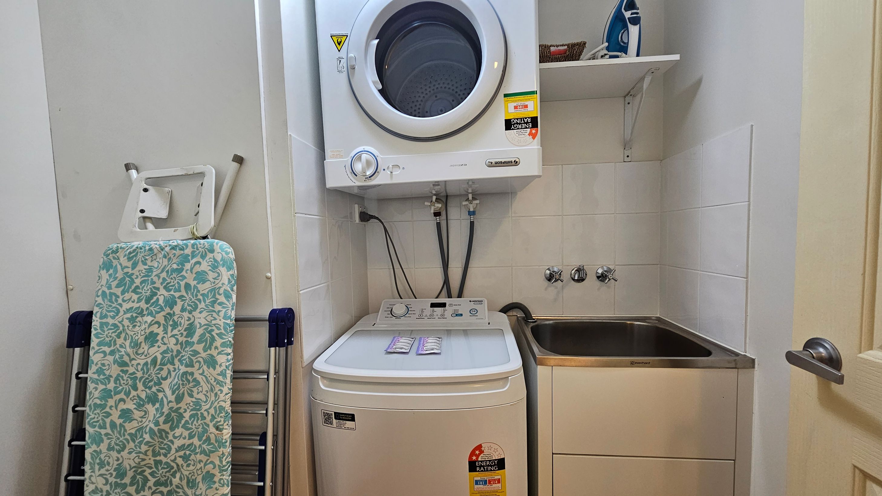 A compact laundry area with a top-loading washing machine and a front-loading dryer stacked above it. To the left, there's a folded ironing board with a floral cover. On the right, a stainless steel sink is set into a cabinet. Above the sink, a shelf holds an iron and a small basket. The walls are tiled, and a door with a silver handle is partially visible on the far right. A compact laundry area with a top-loading washing machine and a front-loading dryer stacked above it. To the left, there's a folded ironing board with a floral cover. On the right, a stainless steel sink is set into a cabinet. Above the sink, a shelf holds an iron and a small basket. The walls are tiled, and a door with a silver handle is partially visible on the far right.