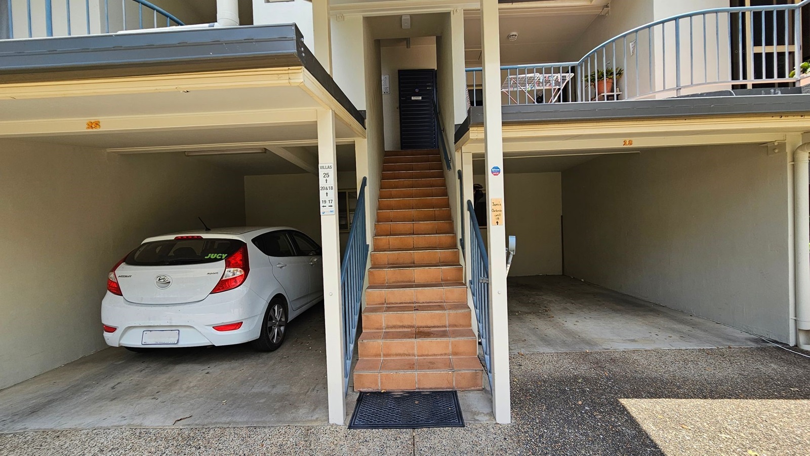 A white Hyundai car is parked in a covered space on the left. In the centre, a set of terracotta-coloured stairs with blue railings leads up to a black door. On the right, there is an empty parking space. Above, a balcony with a blue railing holds a small table and potted plants. The ground is a mix of concrete and gravel. A white Hyundai car is parked in a covered space on the left. In the centre, a set of terracotta-coloured stairs with blue railings leads up to a black door. On the right, there is an empty parking space. Above, a balcony with a blue railing holds a small table and potted plants. The ground is a mix of concrete and gravel.