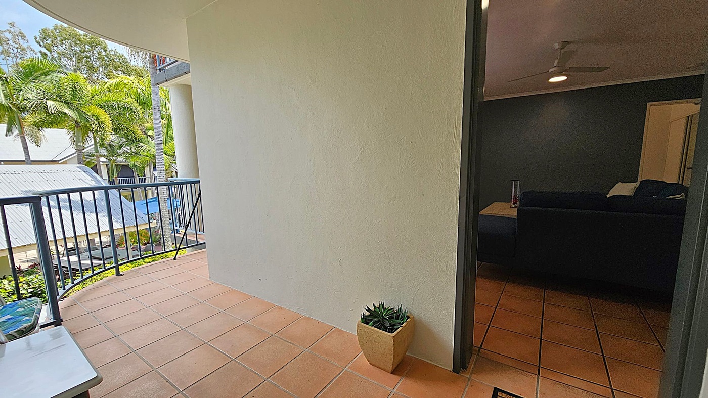 A balcony with terracotta tiles and a metal railing on the left overlooks a garden with palm trees and a building with a corrugated metal roof. A small potted plant sits on the floor near the wall. On the right, an open door leads into a dimly lit room with a dark sofa, a side table, and a ceiling fan. A balcony with terracotta tiles and a metal railing on the left overlooks a garden with palm trees and a building with a corrugated metal roof. A small potted plant sits on the floor near the wall. On the right, an open door leads into a dimly lit room with a dark sofa, a side table, and a ceiling fan.
