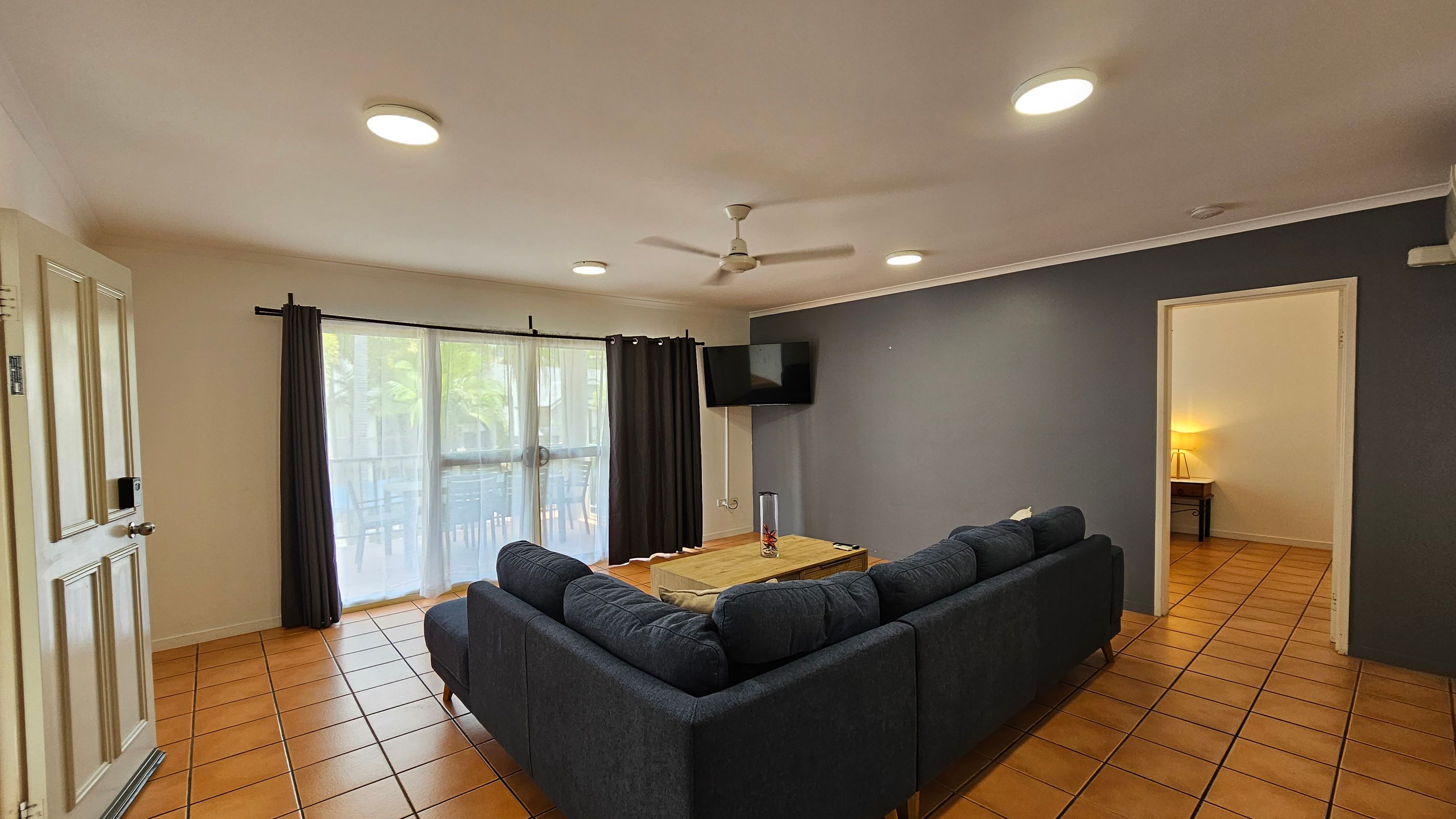 A living room with terracotta tiled flooring and a white ceiling with recessed lights. On the left, there's a partially open door and large windows with sheer and dark curtains. In the centre, a dark grey L-shaped sofa surrounds a wooden coffee table with a decorative vase. A wall-mounted TV is on the right wall, which is painted grey. An open doorway leads to another room with a lit lamp visible. A ceiling fan is centred above the seating area. A living room with terracotta tiled flooring and a white ceiling with recessed lights. On the left, there's a partially open door and large windows with sheer and dark curtains. In the centre, a dark grey L-shaped sofa surrounds a wooden coffee table with a decorative vase. A wall-mounted TV is on the right wall, which is painted grey. An open doorway leads to another room with a lit lamp visible. A ceiling fan is centred above the seating area.