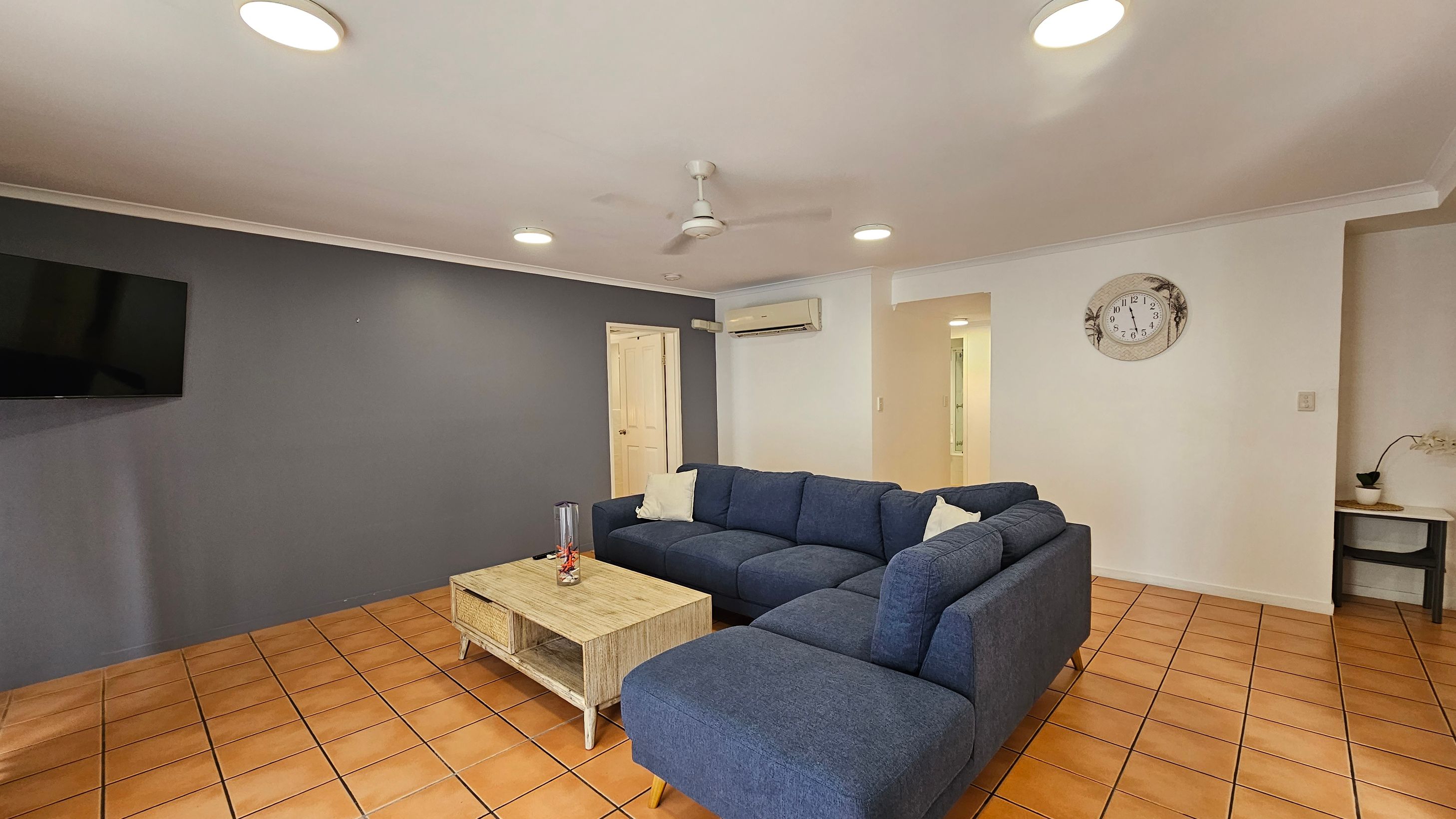 A living room with terracotta tiled flooring and a white ceiling featuring recessed lights and a ceiling fan. On the left, a dark blue accent wall holds a mounted flat-screen TV. In the centre, a large blue L-shaped sofa with white cushions faces a wooden coffee table. On the right, a white wall displays a decorative clock. Below the clock, a small table with a potted plant is visible. An air conditioning unit is mounted above a doorway in the background. A living room with terracotta tiled flooring and a white ceiling featuring recessed lights and a ceiling fan. On the left, a dark blue accent wall holds a mounted flat-screen TV. In the centre, a large blue L-shaped sofa with white cushions faces a wooden coffee table. On the right, a white wall displays a decorative clock. Below the clock, a small table with a potted plant is visible. An air conditioning unit is mounted above a doorway in the background.