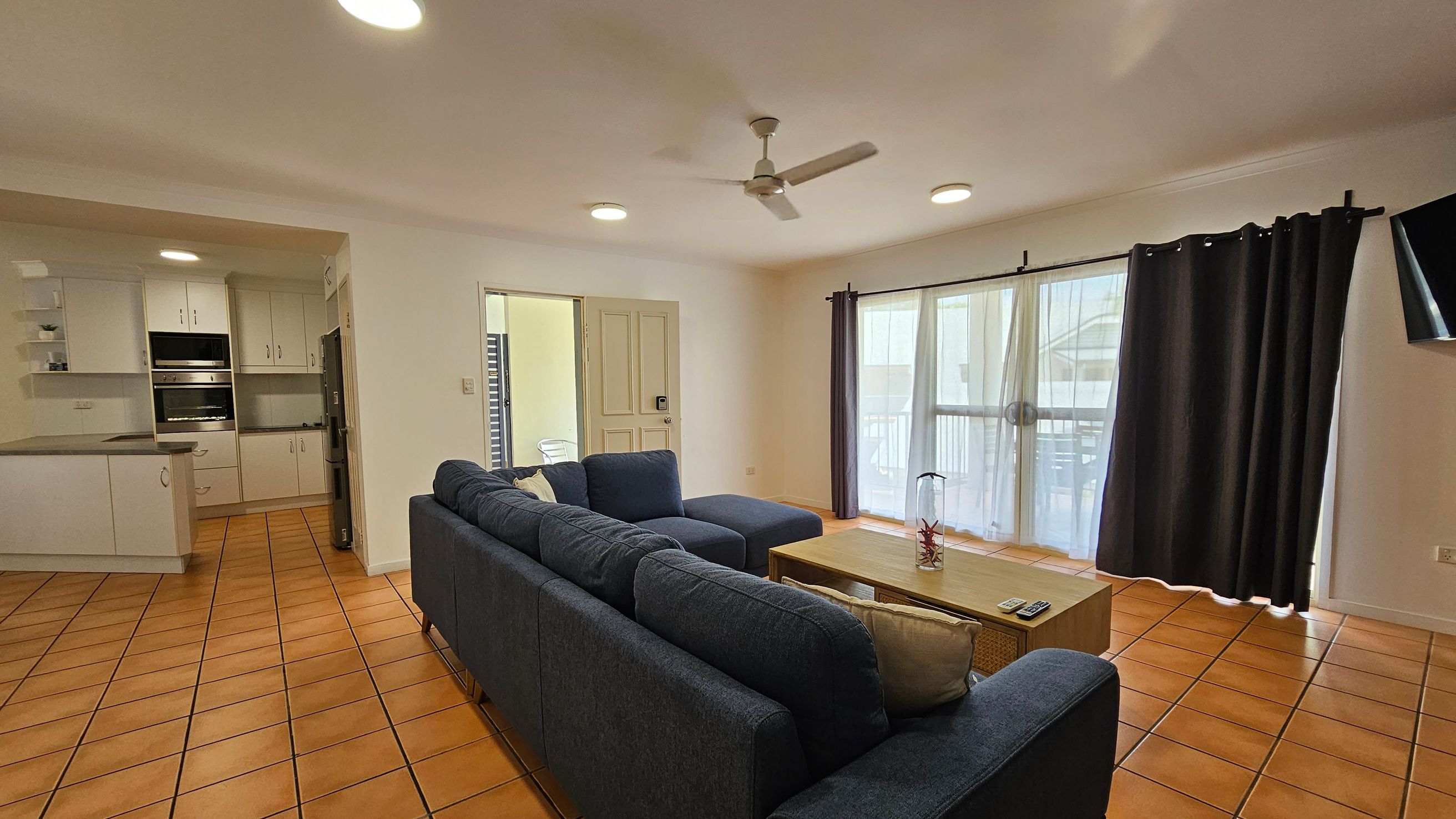 A living room with terracotta tiled flooring and a white ceiling with recessed lights. On the right, a large window with sheer and dark curtains lets in natural light. A dark blue L-shaped sofa faces a wooden coffee table with a decorative item. A flat-screen TV is mounted on the wall. On the left, an open kitchen area features white cabinets, a built-in oven, and a fridge. A ceiling fan is centred above the room. A living room with terracotta tiled flooring and a white ceiling with recessed lights. On the right, a large window with sheer and dark curtains lets in natural light. A dark blue L-shaped sofa faces a wooden coffee table with a decorative item. A flat-screen TV is mounted on the wall. On the left, an open kitchen area features white cabinets, a built-in oven, and a fridge. A ceiling fan is centred above the room.