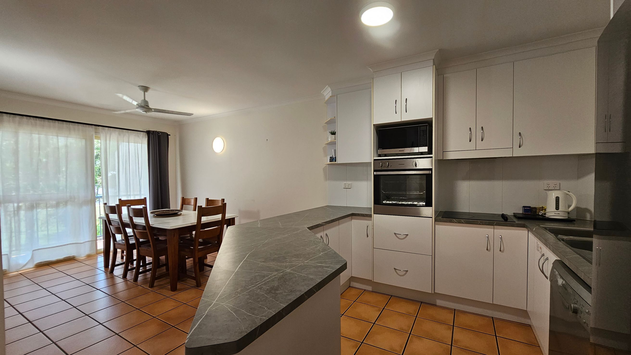 Kitchen and dining area with terracotta floor tiles. On the right, there are white cabinets, a built-in oven, and a kettle on the counter. The kitchen has a dark countertop with a corner sink. On the left, a wooden dining table with six chairs is near large windows covered with sheer curtains. A ceiling fan is above the dining area, and a round wall light is on the wall. Kitchen and dining area with terracotta floor tiles. On the right, there are white cabinets, a built-in oven, and a kettle on the counter. The kitchen has a dark countertop with a corner sink. On the left, a wooden dining table with six chairs is near large windows covered with sheer curtains. A ceiling fan is above the dining area, and a round wall light is on the wall.