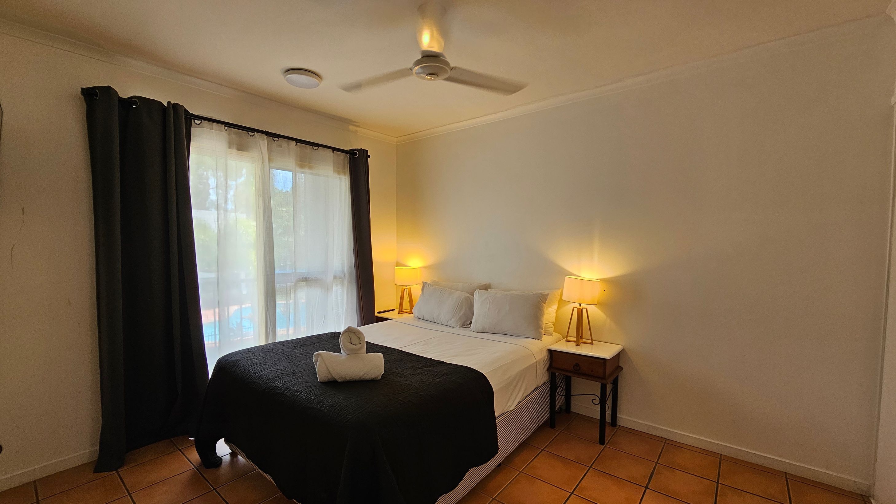 A bedroom with a double bed in the centre, covered with a black quilt and white sheets. Two white pillows are placed at the head of the bed, with a rolled towel on top. On either side of the bed are wooden bedside tables, each with a lit lamp. A ceiling fan is above the bed. To the left, there is a window with sheer white curtains and dark drapes. The floor is tiled in a terracotta colour. A bedroom with a double bed in the centre, covered with a black quilt and white sheets. Two white pillows are placed at the head of the bed, with a rolled towel on top. On either side of the bed are wooden bedside tables, each with a lit lamp. A ceiling fan is above the bed. To the left, there is a window with sheer white curtains and dark drapes. The floor is tiled in a terracotta colour.