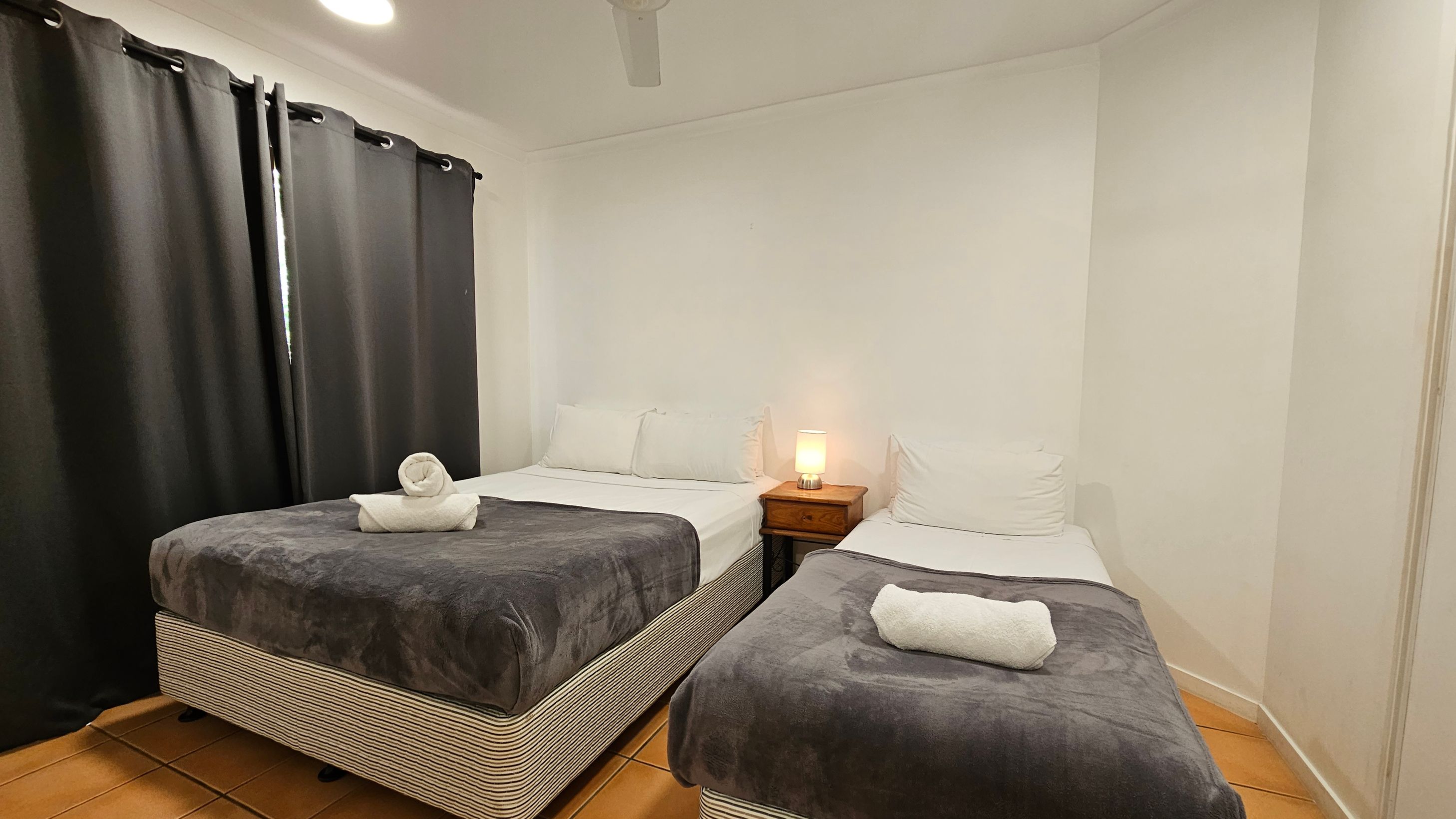 A small bedroom with two single beds, each covered with a dark blanket and a rolled white towel on top. Between the beds is a wooden nightstand with a lit lamp. The left side of the room has dark curtains covering a window. The floor is tiled with a terracotta colour, and the ceiling has a white fan. A small bedroom with two single beds, each covered with a dark blanket and a rolled white towel on top. Between the beds is a wooden nightstand with a lit lamp. The left side of the room has dark curtains covering a window. The floor is tiled with a terracotta colour, and the ceiling has a white fan.