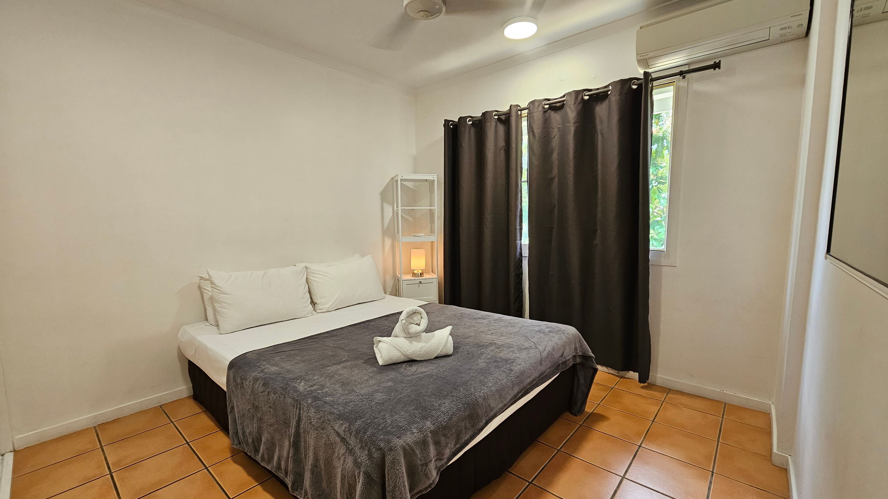 A bedroom with terracotta tiled flooring and a white ceiling. In the centre, there's a double bed with white pillows and a grey blanket, topped with a towel folded into a swan shape. To the right, a window with dark curtains and an air conditioning unit above. A white shelving unit with a lit lamp is next to the bed. A mirror is partially visible on the right wall. A bedroom with terracotta tiled flooring and a white ceiling. In the centre, there's a double bed with white pillows and a grey blanket, topped with a towel folded into a swan shape. To the right, a window with dark curtains and an air conditioning unit above. A white shelving unit with a lit lamp is next to the bed. A mirror is partially visible on the right wall.