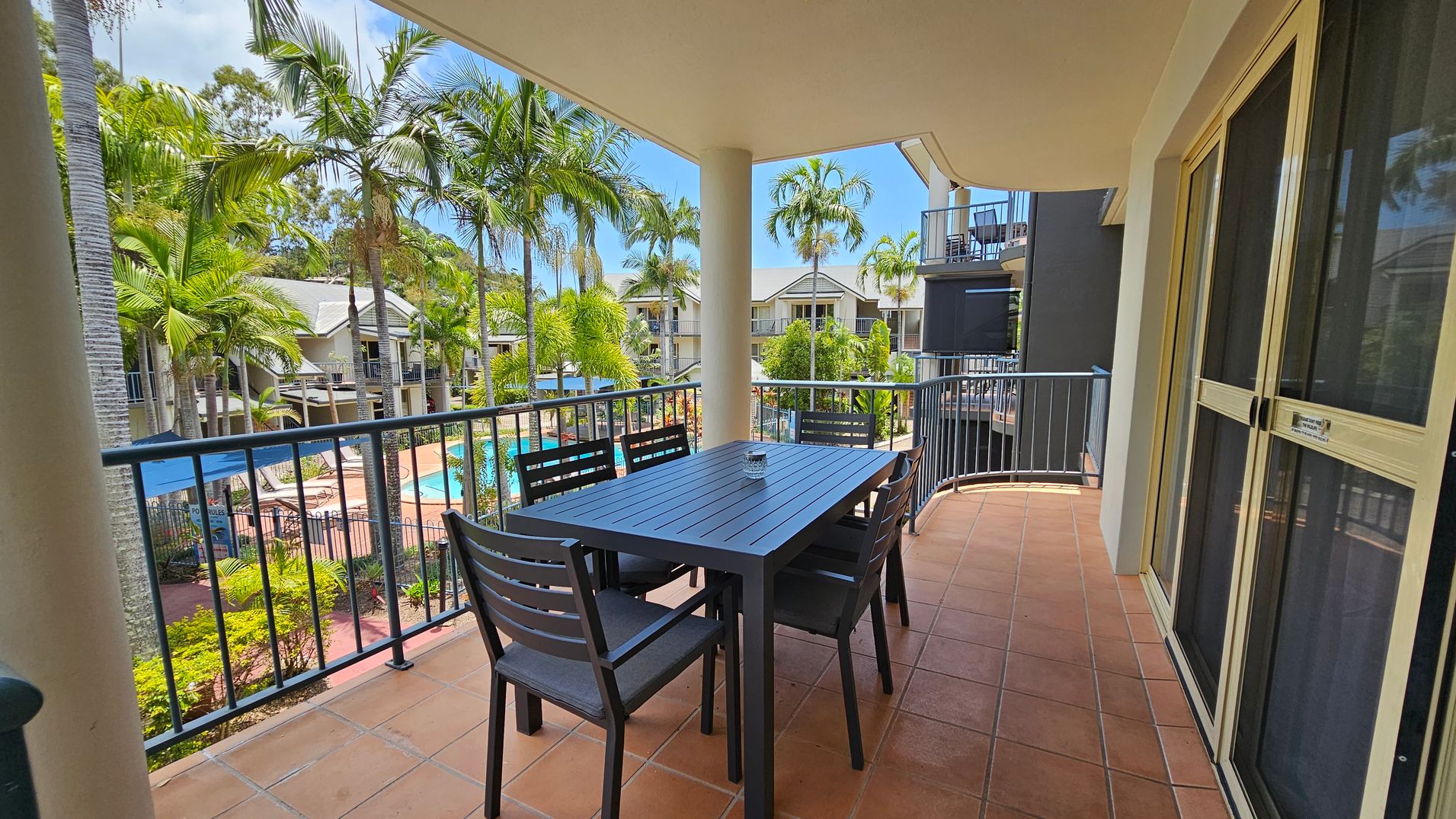 A balcony with terracotta tiles features a dark wooden table and six matching chairs. The railing overlooks a pool area surrounded by palm trees and other greenery. In the background, there are several residential buildings with balconies. On the right, sliding glass doors lead inside. The ceiling is light-coloured and flat. A balcony with terracotta tiles features a dark wooden table and six matching chairs. The railing overlooks a pool area surrounded by palm trees and other greenery. In the background, there are several residential buildings with balconies. On the right, sliding glass doors lead inside. The ceiling is light-coloured and flat.