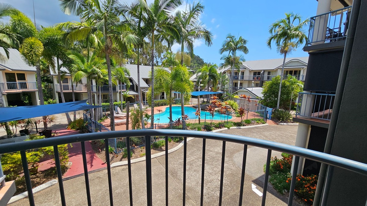 A view from a balcony overlooking a courtyard with a swimming pool surrounded by palm trees. The pool area is fenced and has sun loungers and blue shade sails. There are two-storey buildings with balconies on either side, featuring grey roofs and light-coloured walls. The ground is paved with red and grey paths, and there are landscaped gardens with various plants and flowers. A view from a balcony overlooking a courtyard with a swimming pool surrounded by palm trees. The pool area is fenced and has sun loungers and blue shade sails. There are two-storey buildings with balconies on either side, featuring grey roofs and light-coloured walls. The ground is paved with red and grey paths, and there are landscaped gardens with various plants and flowers.
