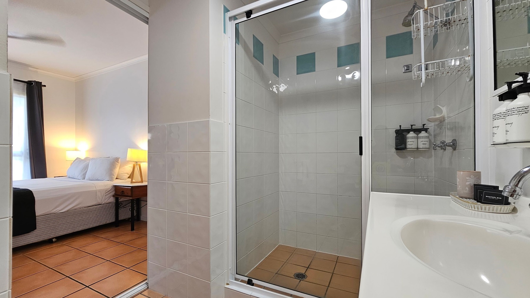 A bathroom with a glass shower enclosure in the centre, featuring white tiles and green accents. Inside the shower, there are wall-mounted toiletries. To the right, a white sink with a chrome tap and various toiletries on a tray. On the left, an open doorway leads to a bedroom with a bed, bedside table, and a lit lamp. The floor is tiled in a terracotta colour. A bathroom with a glass shower enclosure in the centre, featuring white tiles and green accents. Inside the shower, there are wall-mounted toiletries. To the right, a white sink with a chrome tap and various toiletries on a tray. On the left, an open doorway leads to a bedroom with a bed, bedside table, and a lit lamp. The floor is tiled in a terracotta colour.
