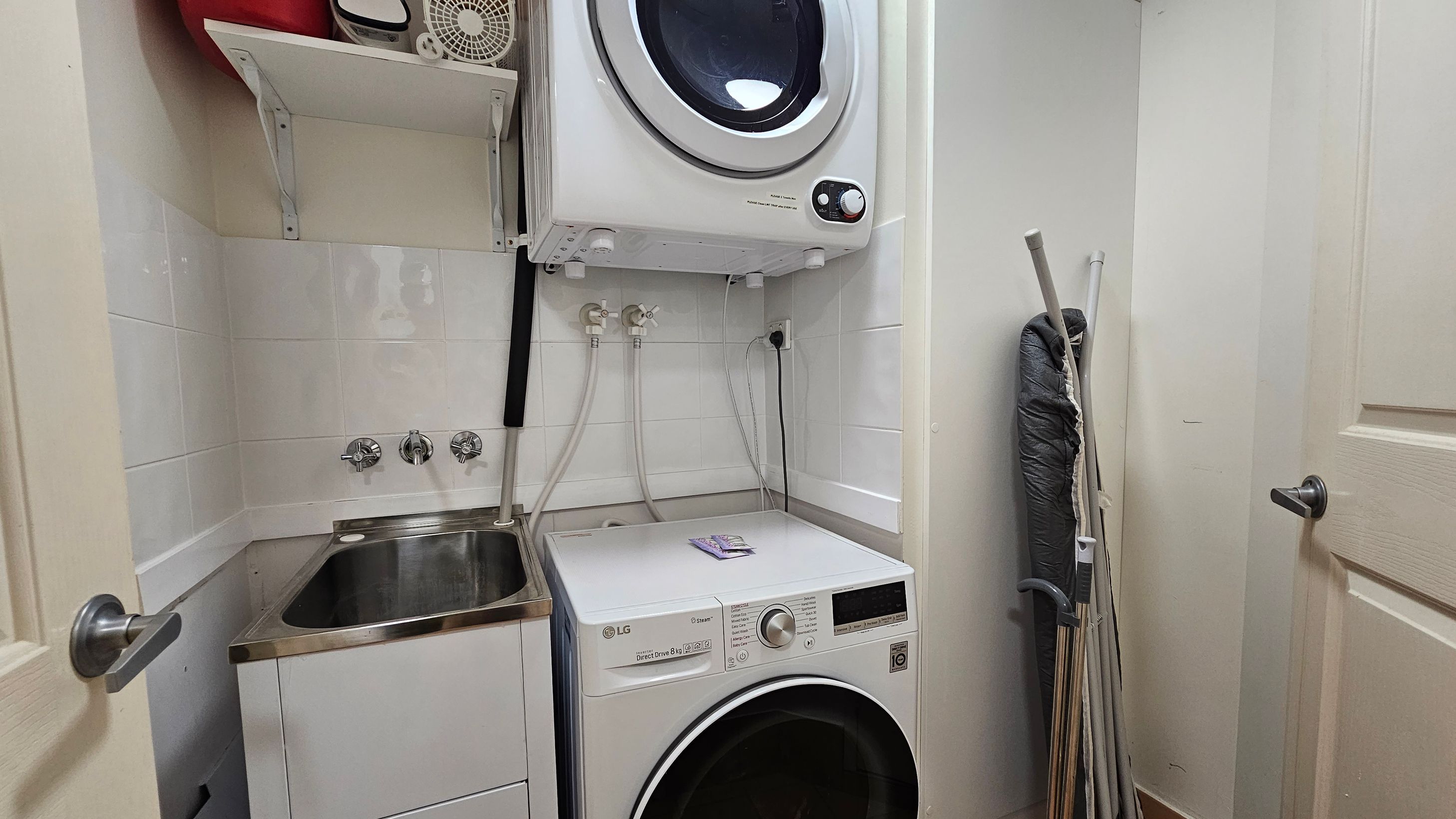 A compact laundry room with a white washing machine on the floor and a dryer stacked above it. To the left, there's a stainless steel sink with taps and a small shelf holding various items, including a fan. On the right, an ironing board and some cleaning tools are propped against the wall. The walls are tiled, and the room is enclosed by a door with a silver handle. A compact laundry room with a white washing machine on the floor and a dryer stacked above it. To the left, there's a stainless steel sink with taps and a small shelf holding various items, including a fan. On the right, an ironing board and some cleaning tools are propped against the wall. The walls are tiled, and the room is enclosed by a door with a silver handle.