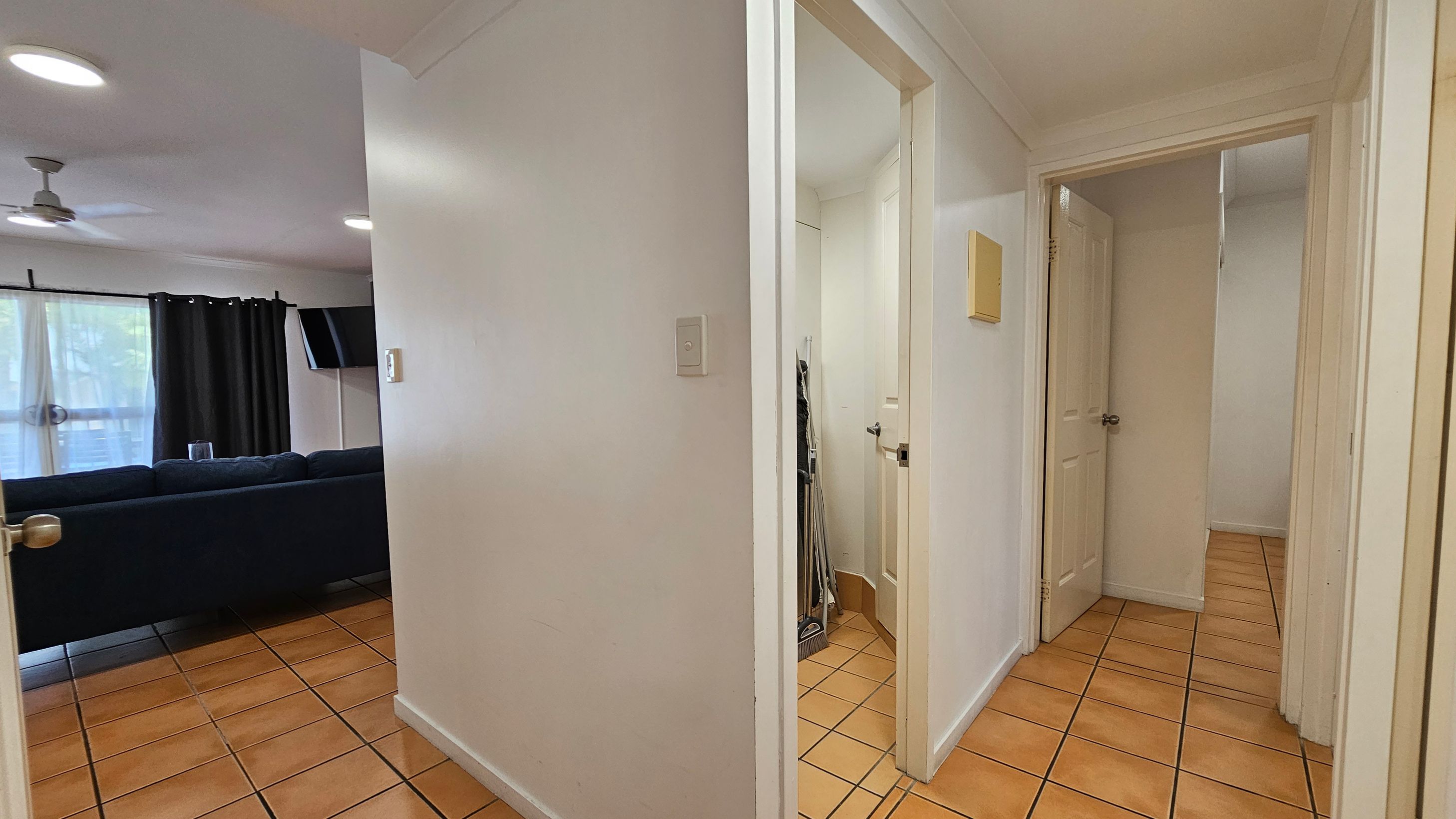 A hallway with terracotta floor tiles and white walls. On the left, an open doorway leads to a living room with a dark sofa, a wall-mounted TV, and a ceiling fan. The room has large windows with dark curtains. On the right, two open doors lead to other rooms, with cleaning tools visible in one doorway. The ceiling is white with recessed lighting. A hallway with terracotta floor tiles and white walls. On the left, an open doorway leads to a living room with a dark sofa, a wall-mounted TV, and a ceiling fan. The room has large windows with dark curtains. On the right, two open doors lead to other rooms, with cleaning tools visible in one doorway. The ceiling is white with recessed lighting.