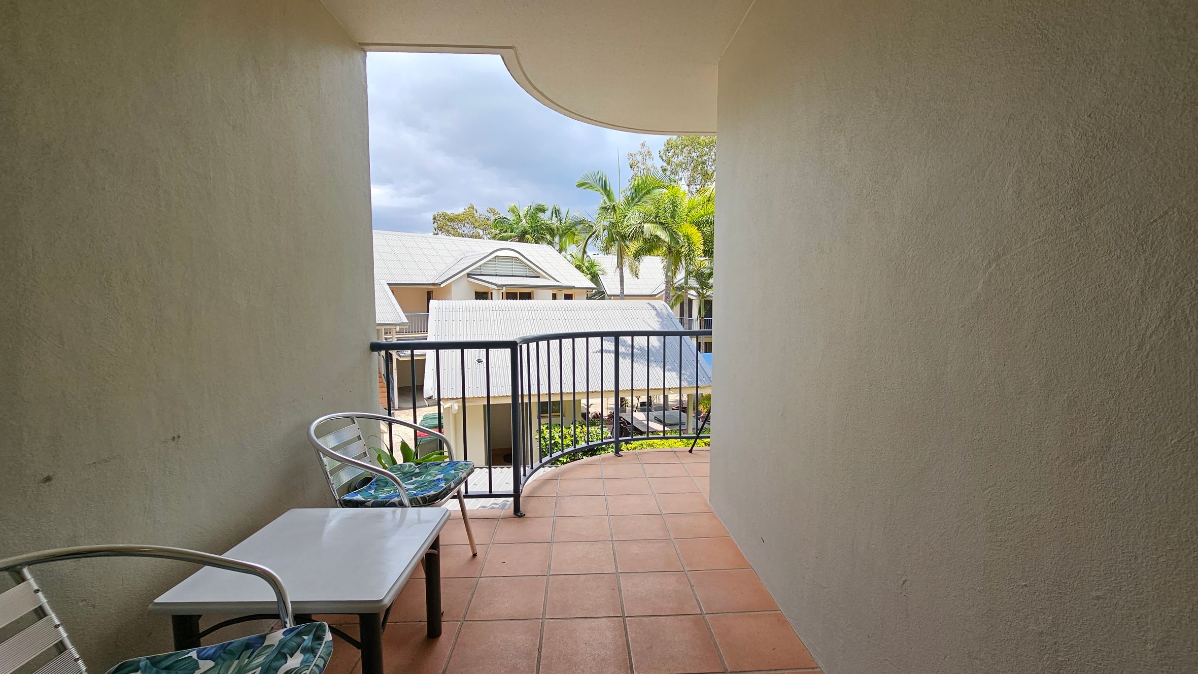 A small balcony with terracotta tiles and a black metal railing. On the left, there are two metal chairs with green patterned cushions and a small white table between them. The view overlooks a complex with light-coloured roofs and palm trees in the background. The walls of the balcony are light-coloured and textured. A small balcony with terracotta tiles and a black metal railing. On the left, there are two metal chairs with green patterned cushions and a small white table between them. The view overlooks a complex with light-coloured roofs and palm trees in the background. The walls of the balcony are light-coloured and textured.