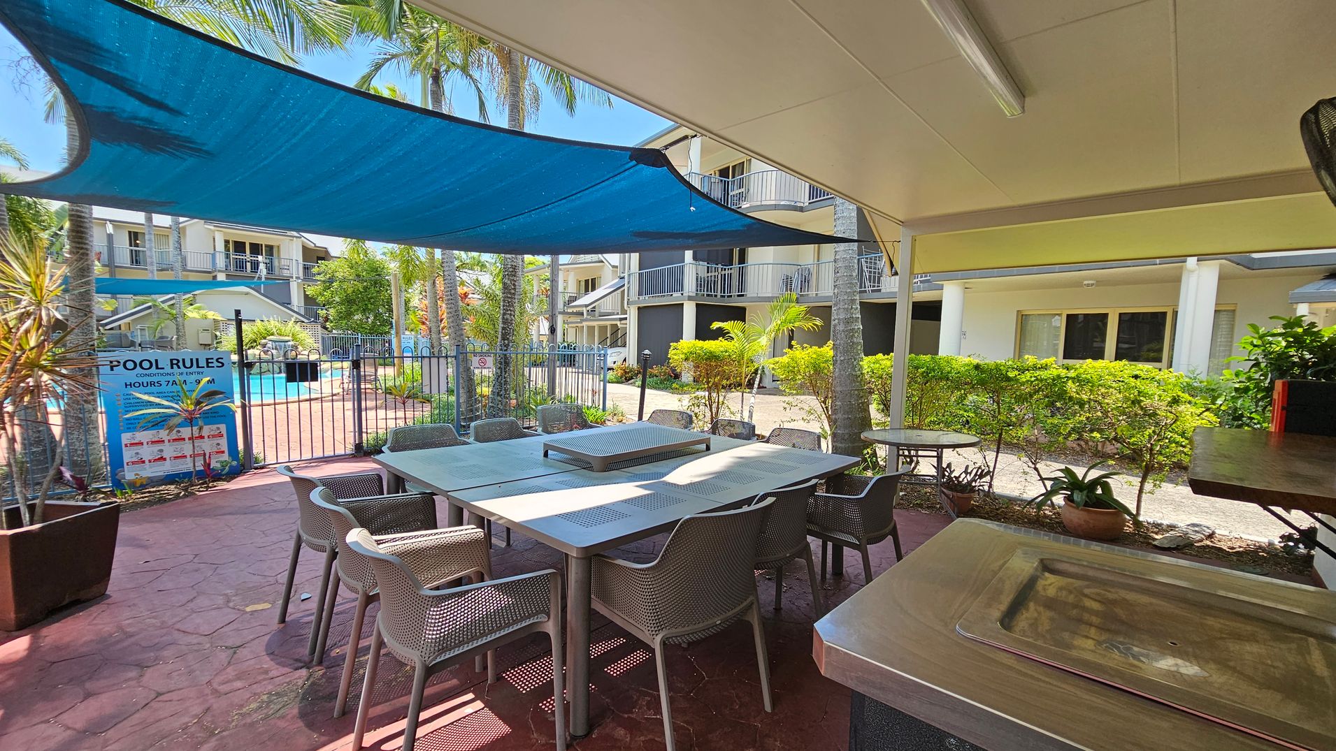 A covered outdoor area with a large metal table surrounded by chairs on a red-tiled floor. To the left, a blue shade sail provides cover, and a sign with pool rules is visible near a fenced swimming pool. In the background, there are multi-storey buildings with balconies, and palm trees and shrubs add greenery to the scene. A metal sink is in the foreground on the right. A covered outdoor area with a large metal table surrounded by chairs on a red-tiled floor. To the left, a blue shade sail provides cover, and a sign with pool rules is visible near a fenced swimming pool. In the background, there are multi-storey buildings with balconies, and palm trees and shrubs add greenery to the scene. A metal sink is in the foreground on the right.