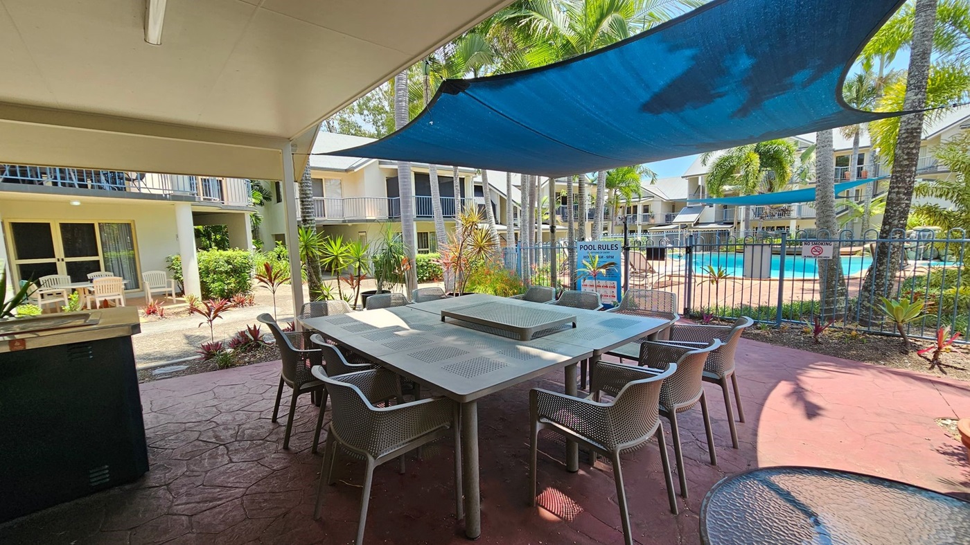 Outdoor seating area with a large square table surrounded by chairs on a red-tiled floor. A blue shade sail is overhead. In the background, there are two-storey buildings with balconies, and a fenced swimming pool area with palm trees and a sign displaying pool rules. White patio chairs are visible near the building entrances, and various plants and shrubs are scattered around the area. Outdoor seating area with a large square table surrounded by chairs on a red-tiled floor. A blue shade sail is overhead. In the background, there are two-storey buildings with balconies, and a fenced swimming pool area with palm trees and a sign displaying pool rules. White patio chairs are visible near the building entrances, and various plants and shrubs are scattered around the area.