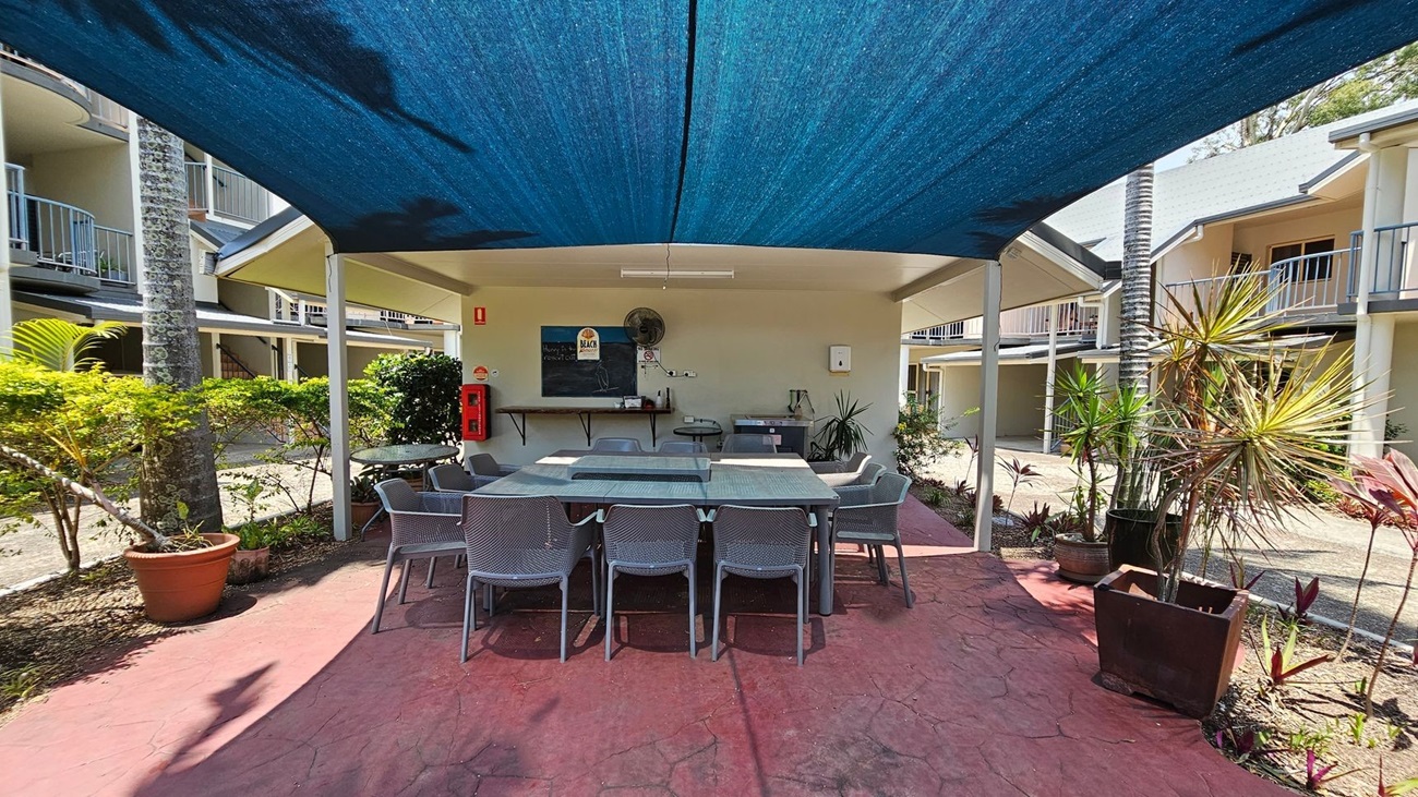 A shaded outdoor area with a large table and several chairs beneath a blue canopy. The ground is paved with red stone. In the background, there's a wall with a chalkboard, a fan, and a fire extinguisher. Potted plants and small trees are on either side. Surrounding the area are two-storey buildings with balconies. A shaded outdoor area with a large table and several chairs beneath a blue canopy. The ground is paved with red stone. In the background, there's a wall with a chalkboard, a fan, and a fire extinguisher. Potted plants and small trees are on either side. Surrounding the area are two-storey buildings with balconies.