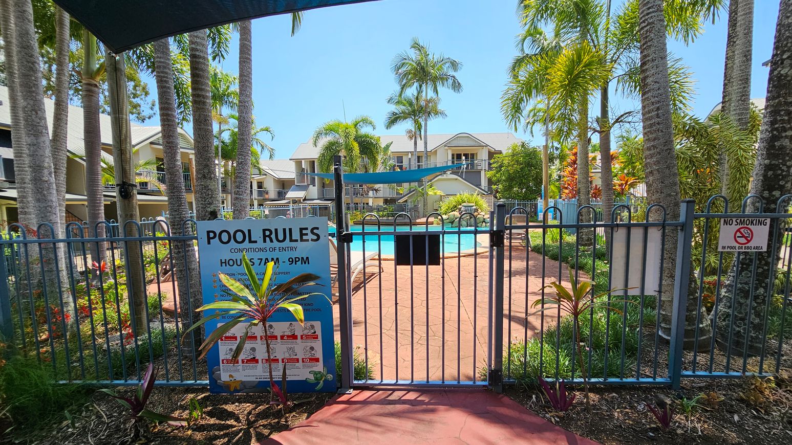 A fenced pool area with a blue gate in the foreground, displaying a "Pool Rules" sign and a "No Smoking" notice. The path leading to the pool is surrounded by lush greenery and tall palm trees. In the background, there are residential buildings with balconies overlooking the pool. The pool is visible, with sun loungers around it, and a shade sail above. The ground is paved with red tiles. A fenced pool area with a blue gate in the foreground, displaying a "Pool Rules" sign and a "No Smoking" notice. The path leading to the pool is surrounded by lush greenery and tall palm trees. In the background, there are residential buildings with balconies overlooking the pool. The pool is visible, with sun loungers around it, and a shade sail above. The ground is paved with red tiles.