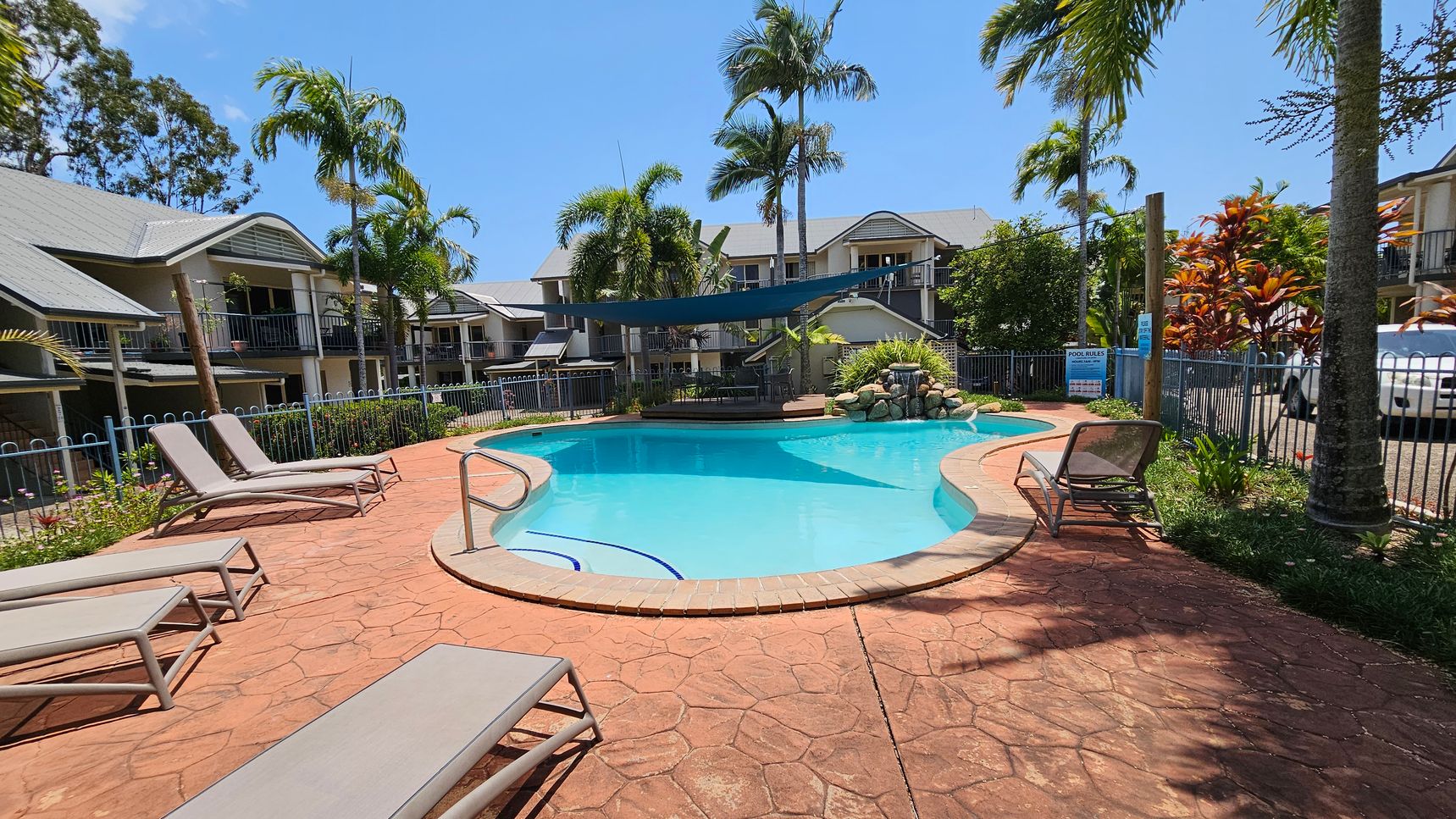 A swimming pool with a curved shape is surrounded by a red-tiled patio. Several sun loungers are placed around the pool. In the background, there are two-storey residential buildings with grey roofs and balconies. Tall palm trees and other tropical plants are scattered around the area. A blue shade sail is stretched over part of the pool, and a small rock waterfall feature is visible at the far end. A white vehicle is parked to the right, near a fence. A swimming pool with a curved shape is surrounded by a red-tiled patio. Several sun loungers are placed around the pool. In the background, there are two-storey residential buildings with grey roofs and balconies. Tall palm trees and other tropical plants are scattered around the area. A blue shade sail is stretched over part of the pool, and a small rock waterfall feature is visible at the far end. A white vehicle is parked to the right, near a fence.