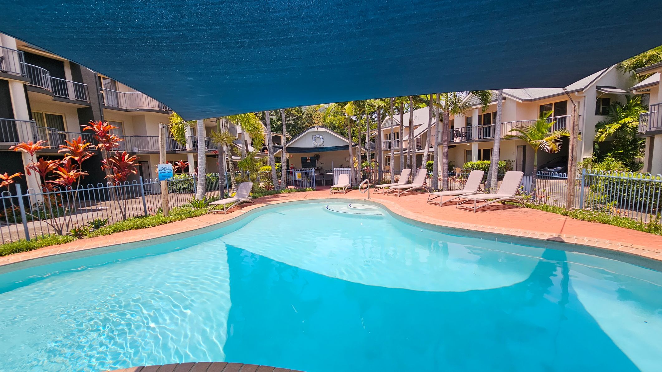 A swimming pool with clear blue water is in the foreground, surrounded by a red brick deck. Several sun loungers are placed along the right side. In the background, there are two-storey buildings with balconies, and a small structure with a clock. Tall palm trees and red-leaved plants are scattered around the area. A blue shade cloth is stretched above the pool, providing partial cover. A metal fence encloses the pool area. A swimming pool with clear blue water is in the foreground, surrounded by a red brick deck. Several sun loungers are placed along the right side. In the background, there are two-storey buildings with balconies, and a small structure with a clock. Tall palm trees and red-leaved plants are scattered around the area. A blue shade cloth is stretched above the pool, providing partial cover. A metal fence encloses the pool area.
