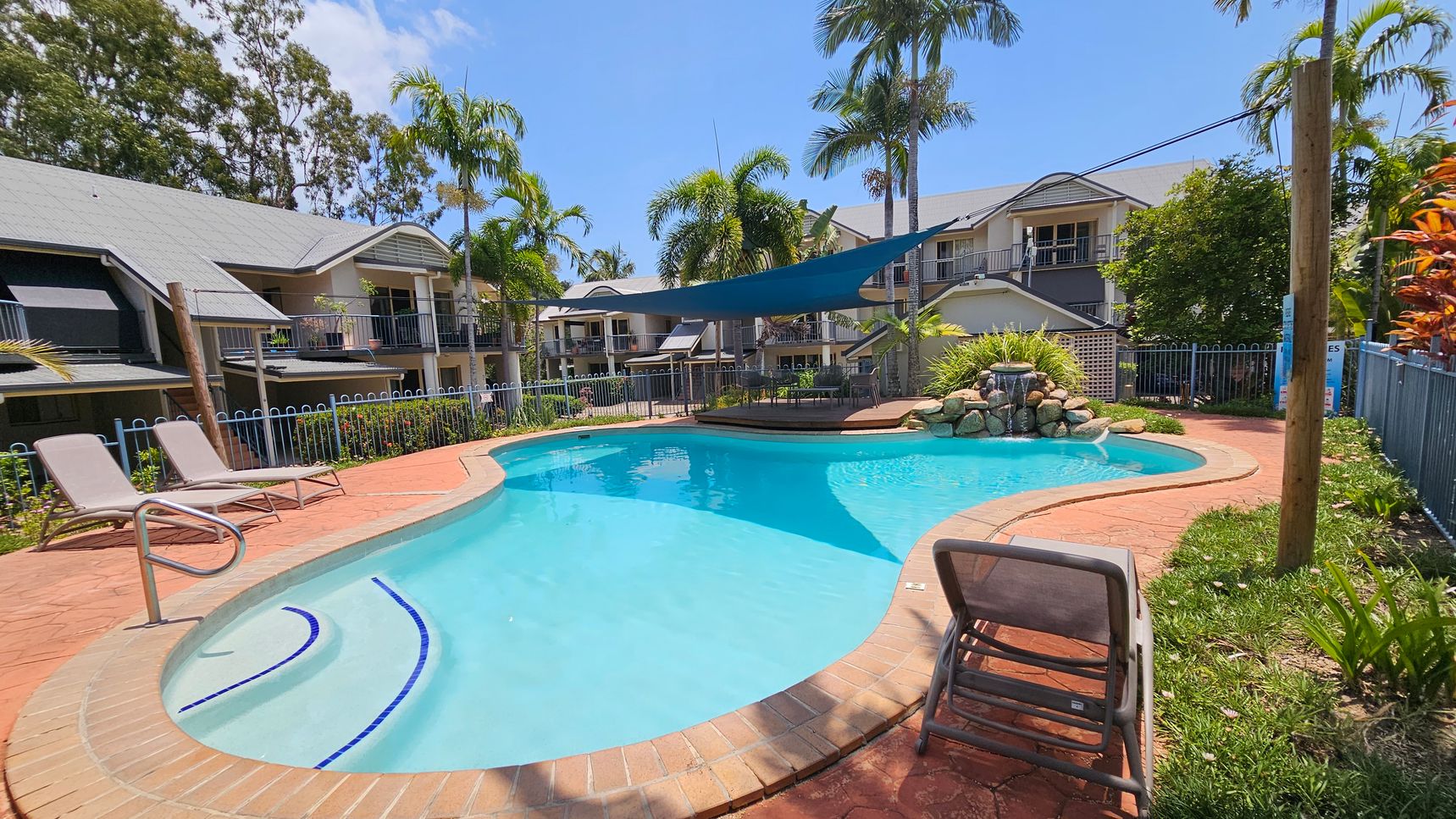 A swimming pool with clear blue water is surrounded by a red brick patio. On the left, two sun loungers are positioned near the pool's edge. In the background, there are several two-storey buildings with grey roofs and balconies. Palm trees and other greenery are scattered around the area. A blue shade sail is stretched over part of the pool, and a small waterfall feature is visible at the far end. A fence encloses the pool area. A swimming pool with clear blue water is surrounded by a red brick patio. On the left, two sun loungers are positioned near the pool's edge. In the background, there are several two-storey buildings with grey roofs and balconies. Palm trees and other greenery are scattered around the area. A blue shade sail is stretched over part of the pool, and a small waterfall feature is visible at the far end. A fence encloses the pool area.