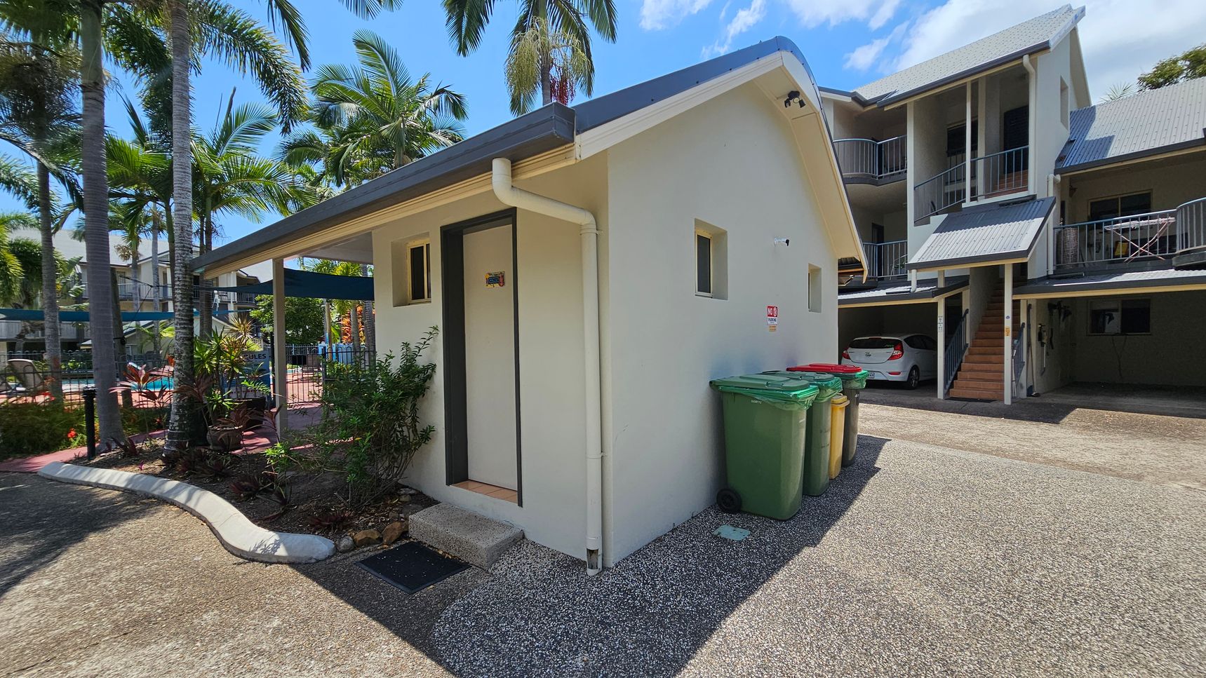 A small, light-coloured building with a sloped roof is in the foreground, featuring a door and two small windows. Green and yellow bins are placed against its side. To the right, a larger building with a grey roof and balconies is visible, with a white car parked underneath. On the left, there are palm trees and a glimpse of a swimming pool area with a fence and sun loungers. The ground is a mix of concrete and gravel. A small, light-coloured building with a sloped roof is in the foreground, featuring a door and two small windows. Green and yellow bins are placed against its side. To the right, a larger building with a grey roof and balconies is visible, with a white car parked underneath. On the left, there are palm trees and a glimpse of a swimming pool area with a fence and sun loungers. The ground is a mix of concrete and gravel.