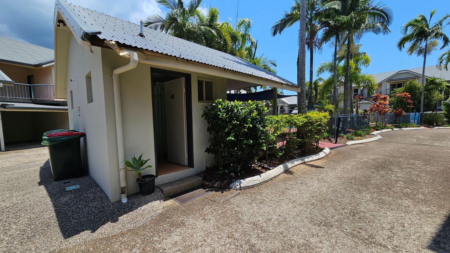 A small building with a corrugated metal roof is on the left, featuring a partially open door and two small windows. A black bin with a red lid and a green bin are beside the building. A potted plant sits near the entrance. On the right, a pathway lined with lush green bushes and tall palm trees leads to a residential area with houses in the background. The ground is covered with gravel, and the area is well-lit by sunlight. A small building with a corrugated metal roof is on the left, featuring a partially open door and two small windows. A black bin with a red lid and a green bin are beside the building. A potted plant sits near the entrance. On the right, a pathway lined with lush green bushes and tall palm trees leads to a residential area with houses in the background. The ground is covered with gravel, and the area is well-lit by sunlight.