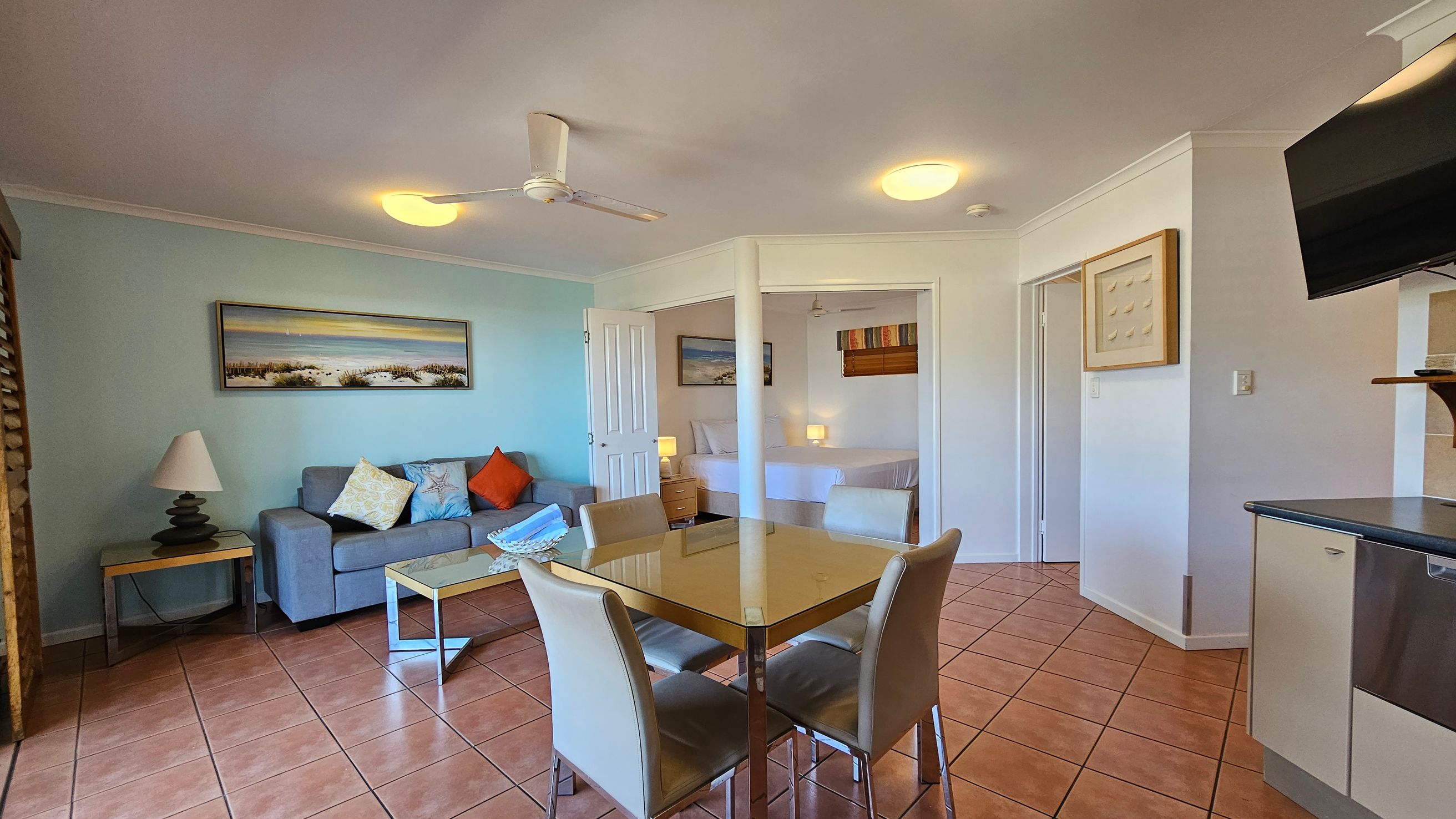 A living room with terracotta tiled flooring and a ceiling fan. On the left, there are wooden shutters leading to a balcony with a view of green hills. In the centre, a glass dining table with four chairs is placed. On the right, a grey sofa with colourful cushions sits against a light blue wall, above which hangs a landscape painting. A glass coffee table is in front of the sofa. A small wooden cabinet with a lamp is near a white door. A living room with terracotta tiled flooring and a ceiling fan. On the left, there are wooden shutters leading to a balcony with a view of green hills. In the centre, a glass dining table with four chairs is placed. On the right, a grey sofa with colourful cushions sits against a light blue wall, above which hangs a landscape painting. A glass coffee table is in front of the sofa. A small wooden cabinet with a lamp is near a white door.