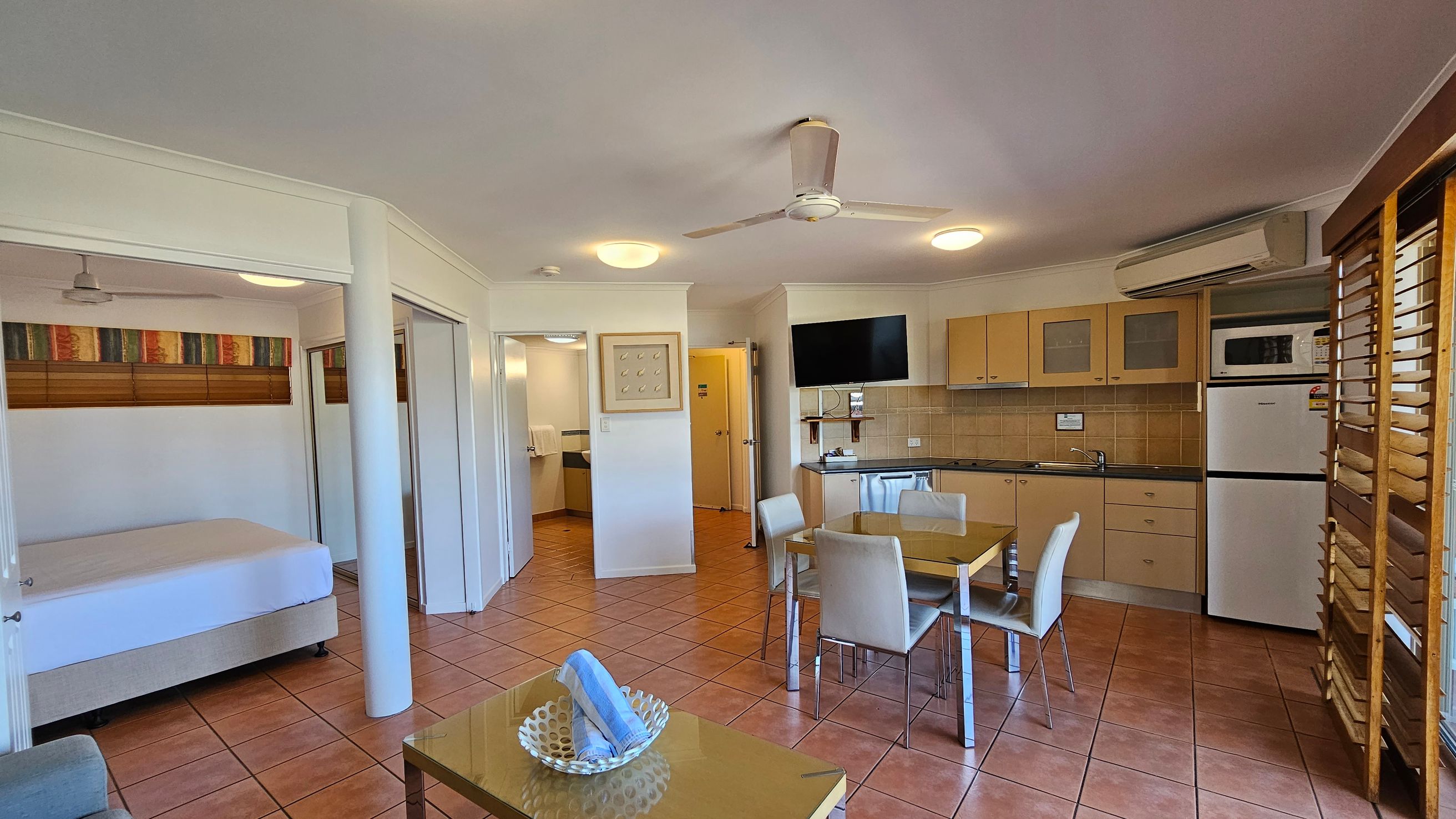 A room with terracotta tiled flooring and a white ceiling with a ceiling fan. On the left, there's a bed with a colourful headboard and a ceiling fan above. In the centre, a dining table with four chairs is placed near a kitchenette with light wood cabinets, a microwave, and a fridge. A TV is mounted on the wall above the kitchen counter. On the right, wooden louvres cover the windows. A small table with a decorative bowl and rolled towels is in the foreground. A room with terracotta tiled flooring and a white ceiling with a ceiling fan. On the left, there's a bed with a colourful headboard and a ceiling fan above. In the centre, a dining table with four chairs is placed near a kitchenette with light wood cabinets, a microwave, and a fridge. A TV is mounted on the wall above the kitchen counter. On the right, wooden louvres cover the windows. A small table with a decorative bowl and rolled towels is in the foreground.