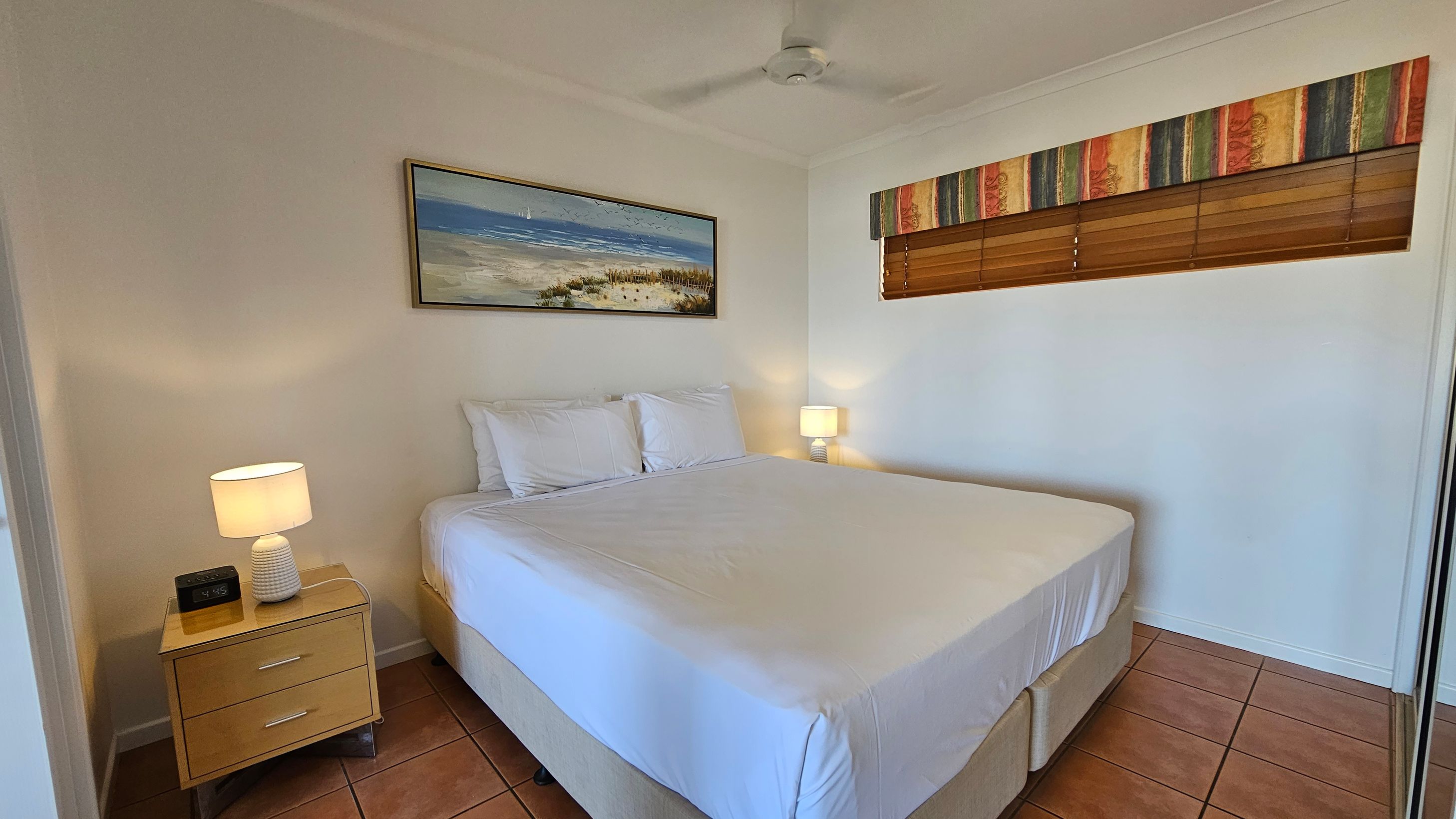 A bedroom with a large bed in the centre, covered in white linens and pillows. On the left, a wooden bedside table holds a lamp and a digital clock. Above the bed, a framed beach scene painting hangs on the wall. On the right, a window with wooden blinds and a colourful valance. The floor is tiled in a terracotta colour, and a ceiling fan is visible above. A bedroom with a large bed in the centre, covered in white linens and pillows. On the left, a wooden bedside table holds a lamp and a digital clock. Above the bed, a framed beach scene painting hangs on the wall. On the right, a window with wooden blinds and a colourful valance. The floor is tiled in a terracotta colour, and a ceiling fan is visible above.