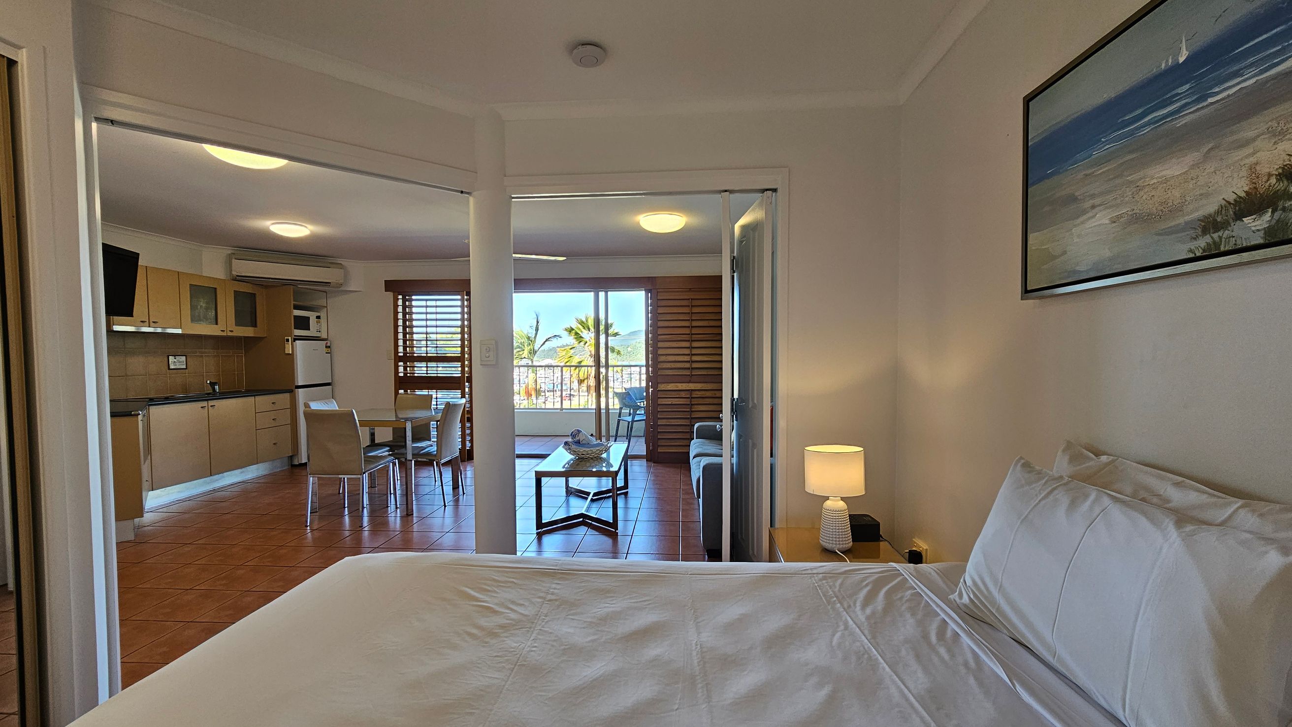 A bedroom with a neatly made bed and a bedside table with a lamp on the right. A framed picture hangs above the bed. Through an open doorway, a living area is visible with a kitchen on the left, featuring light wood cabinets and a small dining table with chairs. Beyond, a sitting area with a sofa and coffee table is seen, leading to a balcony with a view of palm trees. The floor is tiled, and the ceiling has recessed lighting. A bedroom with a neatly made bed and a bedside table with a lamp on the right. A framed picture hangs above the bed. Through an open doorway, a living area is visible with a kitchen on the left, featuring light wood cabinets and a small dining table with chairs. Beyond, a sitting area with a sofa and coffee table is seen, leading to a balcony with a view of palm trees. The floor is tiled, and the ceiling has recessed lighting.