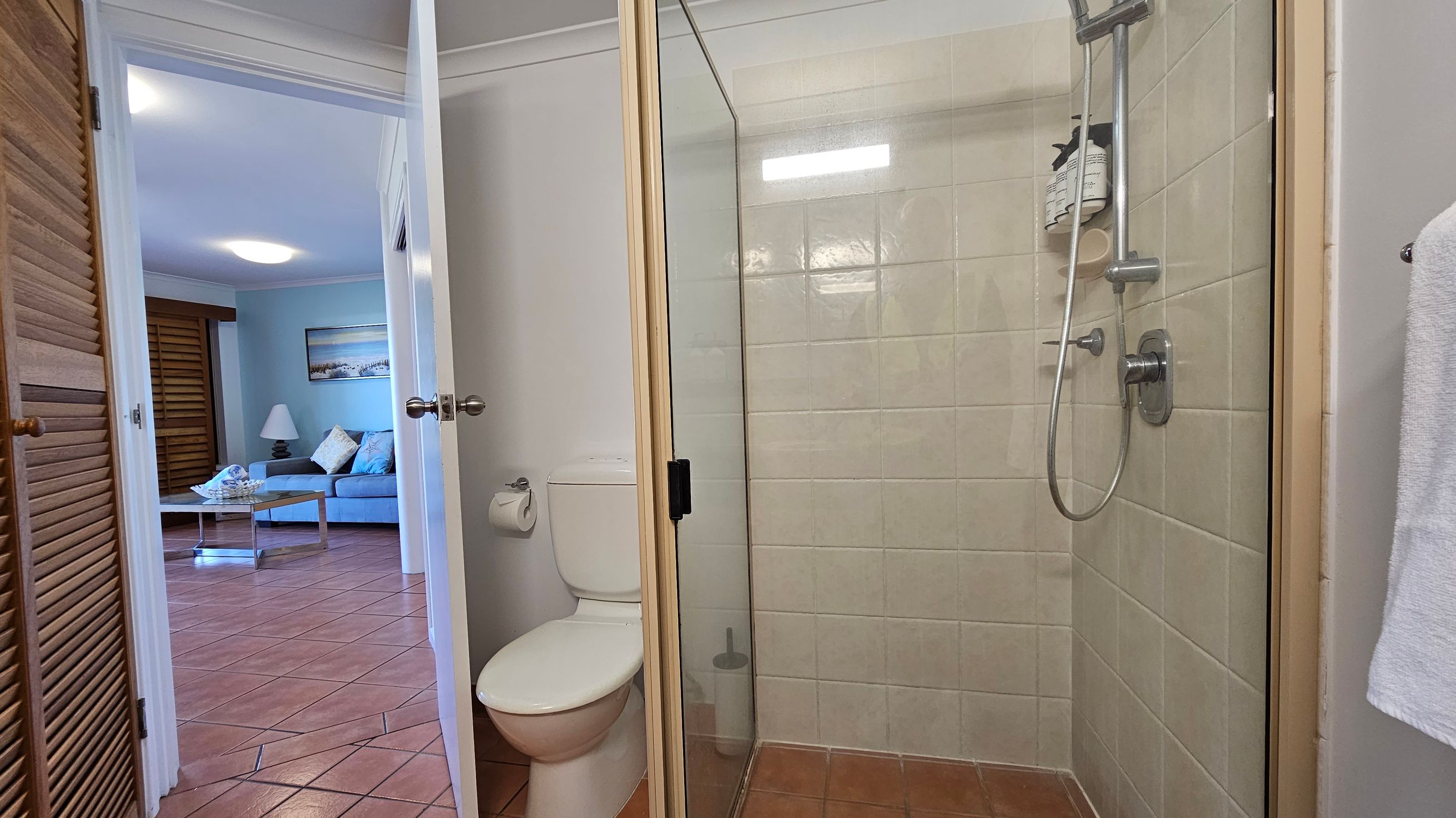 A bathroom with a glass shower enclosure on the right, featuring beige tiles and a silver showerhead. A white toilet is next to the shower, with a toilet roll holder on the wall. The door is open, leading to a room with terracotta floor tiles. In the adjacent room, a blue sofa, a lamp, and a coffee table are visible. A painting hangs on the wall, and wooden shutters cover a window. A bathroom with a glass shower enclosure on the right, featuring beige tiles and a silver showerhead. A white toilet is next to the shower, with a toilet roll holder on the wall. The door is open, leading to a room with terracotta floor tiles. In the adjacent room, a blue sofa, a lamp, and a coffee table are visible. A painting hangs on the wall, and wooden shutters cover a window.