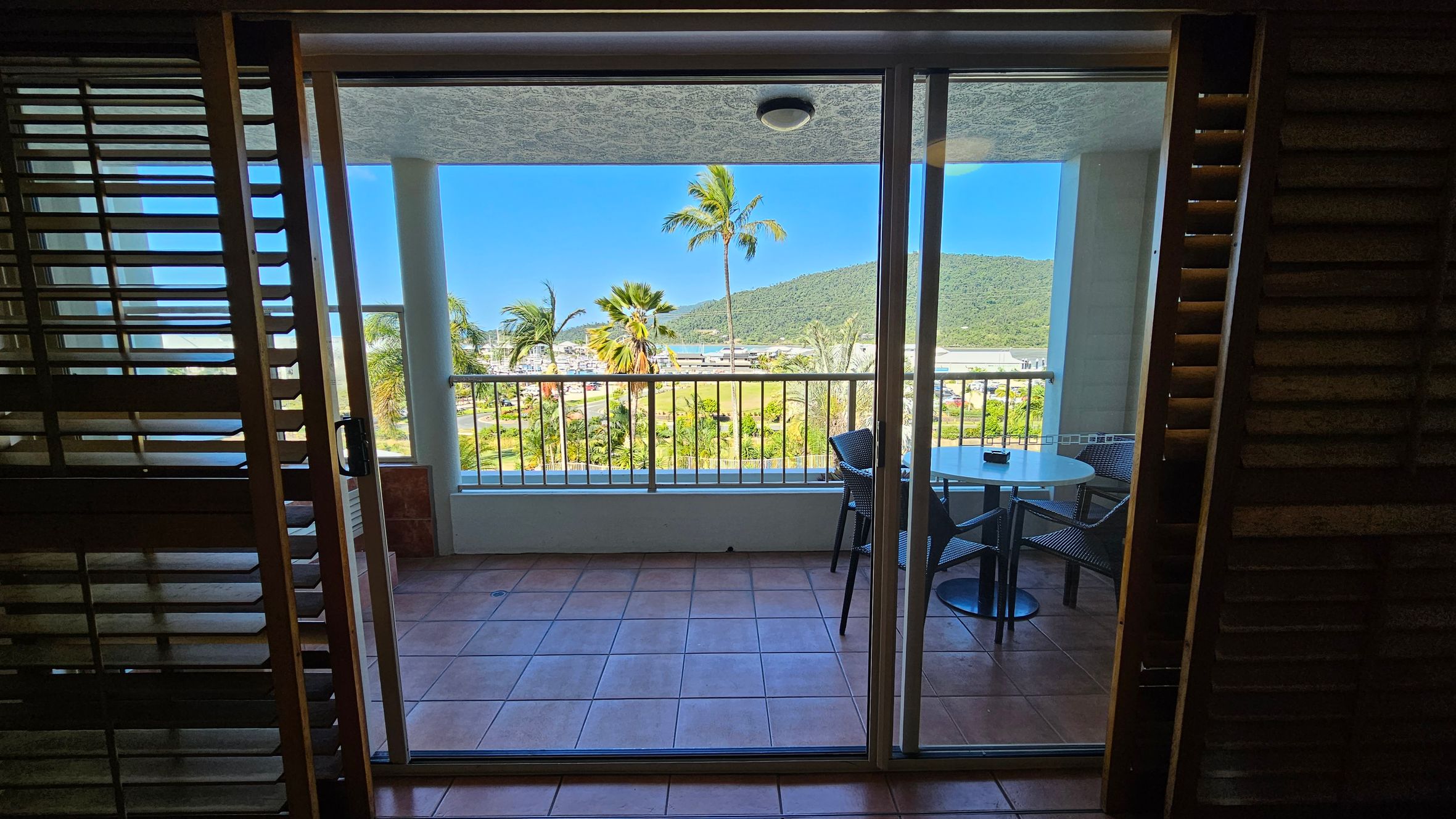 A view through a glass sliding door leading to a balcony with terracotta tiles. On the balcony, there's a round table with two chairs. Beyond the railing, palm trees and lush greenery are visible, with a hill in the background. The ceiling of the balcony is white with a light fixture. Wooden shutters are partially open on either side of the door. A view through a glass sliding door leading to a balcony with terracotta tiles. On the balcony, there's a round table with two chairs. Beyond the railing, palm trees and lush greenery are visible, with a hill in the background. The ceiling of the balcony is white with a light fixture. Wooden shutters are partially open on either side of the door.