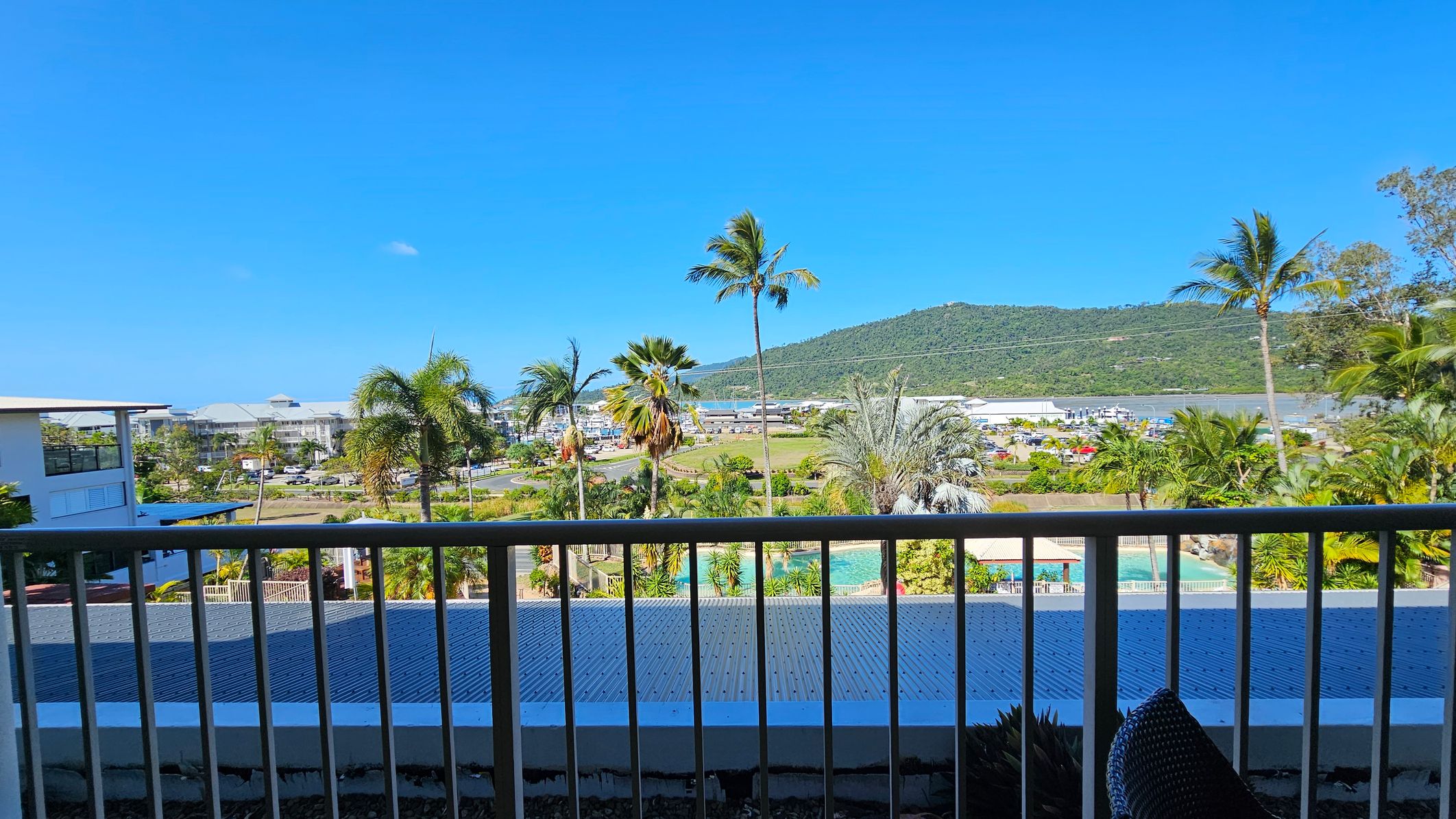 A view from a balcony with a railing in the foreground. Below, there's a corrugated metal roof and a swimming pool surrounded by lush greenery and palm trees. In the background, a small town with buildings and parked cars is visible. Beyond the town, a green hill rises under a clear blue sky. A view from a balcony with a railing in the foreground. Below, there's a corrugated metal roof and a swimming pool surrounded by lush greenery and palm trees. In the background, a small town with buildings and parked cars is visible. Beyond the town, a green hill rises under a clear blue sky.