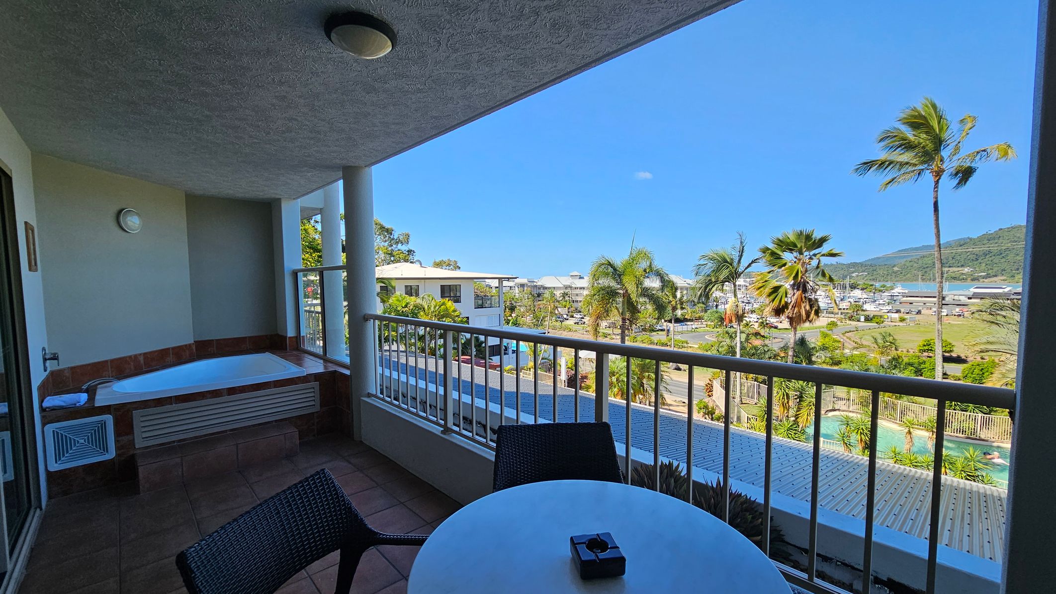 A balcony with a tiled floor features a round table and two chairs. On the left, there's a built-in bathtub with a towel. The railing overlooks a view of palm trees, a swimming pool, and a marina in the distance. The ceiling has a light fixture. The background includes buildings and a hillside. A balcony with a tiled floor features a round table and two chairs. On the left, there's a built-in bathtub with a towel. The railing overlooks a view of palm trees, a swimming pool, and a marina in the distance. The ceiling has a light fixture. The background includes buildings and a hillside.