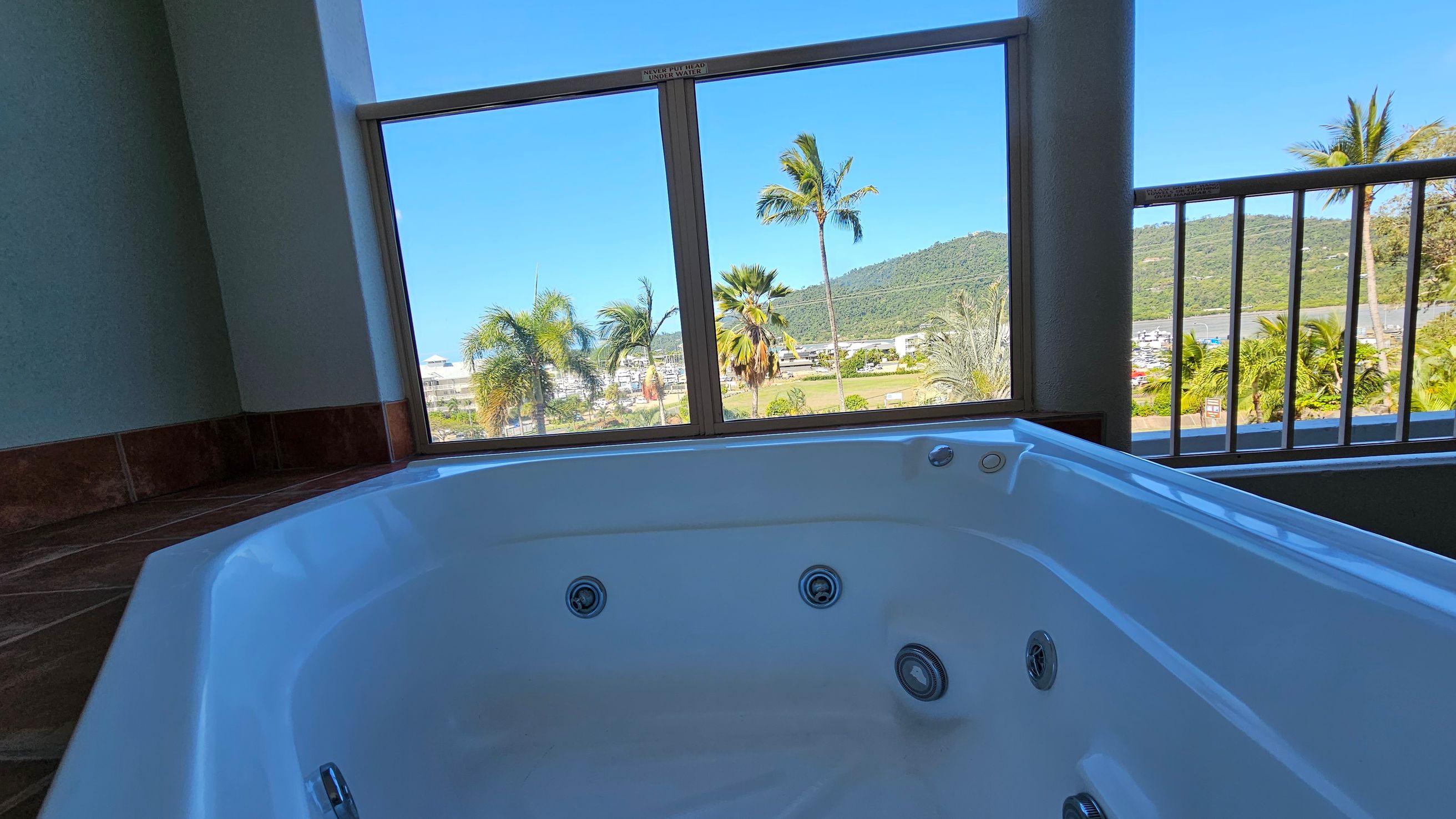 A white jacuzzi tub is in the foreground, positioned indoors near large windows. The windows offer a view of palm trees, a grassy area, and a distant hill. The floor is tiled with dark tiles, and the ceiling is not visible. The scene outside is bright and sunny, with clear blue skies. A white jacuzzi tub is in the foreground, positioned indoors near large windows. The windows offer a view of palm trees, a grassy area, and a distant hill. The floor is tiled with dark tiles, and the ceiling is not visible. The scene outside is bright and sunny, with clear blue skies.
