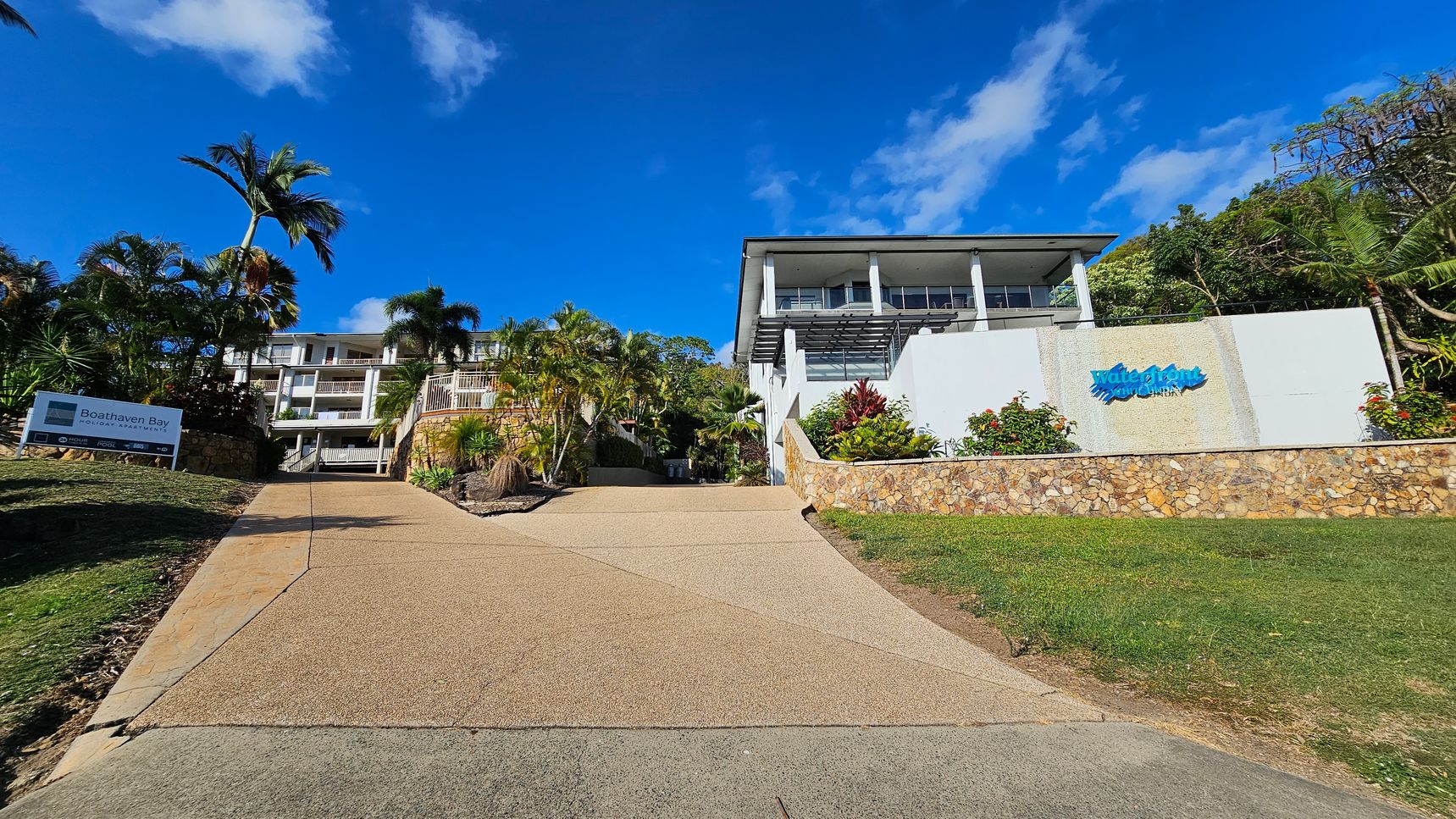 A paved driveway leads up to a multi-storey building surrounded by lush palm trees and tropical plants. On the left, a sign reads "Boathaven Bay Holiday Apartments." The building features balconies and is set against a backdrop of clear blue sky. The foreground includes a grassy slope with scattered rocks and a variety of greenery. A paved driveway leads up to a multi-storey building surrounded by lush palm trees and tropical plants. On the left, a sign reads "Boathaven Bay Holiday Apartments." The building features balconies and is set against a backdrop of clear blue sky. The foreground includes a grassy slope with scattered rocks and a variety of greenery.