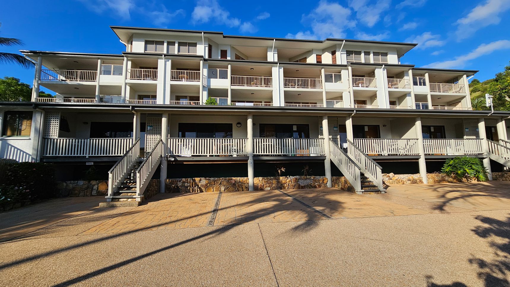 A multi-storey building with a series of balconies and white railings. The structure has a light green facade and is elevated on stone foundations. Several staircases lead up to the main entrance, which is shaded by a roof supported by columns. The foreground features a paved area with shadows of palm trees. The sky is blue with some clouds. A multi-storey building with a series of balconies and white railings. The structure has a light green facade and is elevated on stone foundations. Several staircases lead up to the main entrance, which is shaded by a roof supported by columns. The foreground features a paved area with shadows of palm trees. The sky is blue with some clouds.