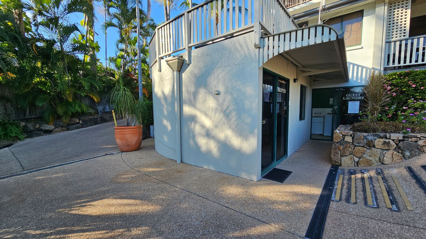 A curved building corner with a glass door labelled "Reception" on the right. Next to it, a sign reads "Guest Laundry" above a stone planter with pink flowers. A large terracotta pot with a tall plant is on the left, near a sloped path lined with lush green palm trees. The ground is textured concrete, and a metal railing runs along the top of the building. A curved building corner with a glass door labelled "Reception" on the right. Next to it, a sign reads "Guest Laundry" above a stone planter with pink flowers. A large terracotta pot with a tall plant is on the left, near a sloped path lined with lush green palm trees. The ground is textured concrete, and a metal railing runs along the top of the building.