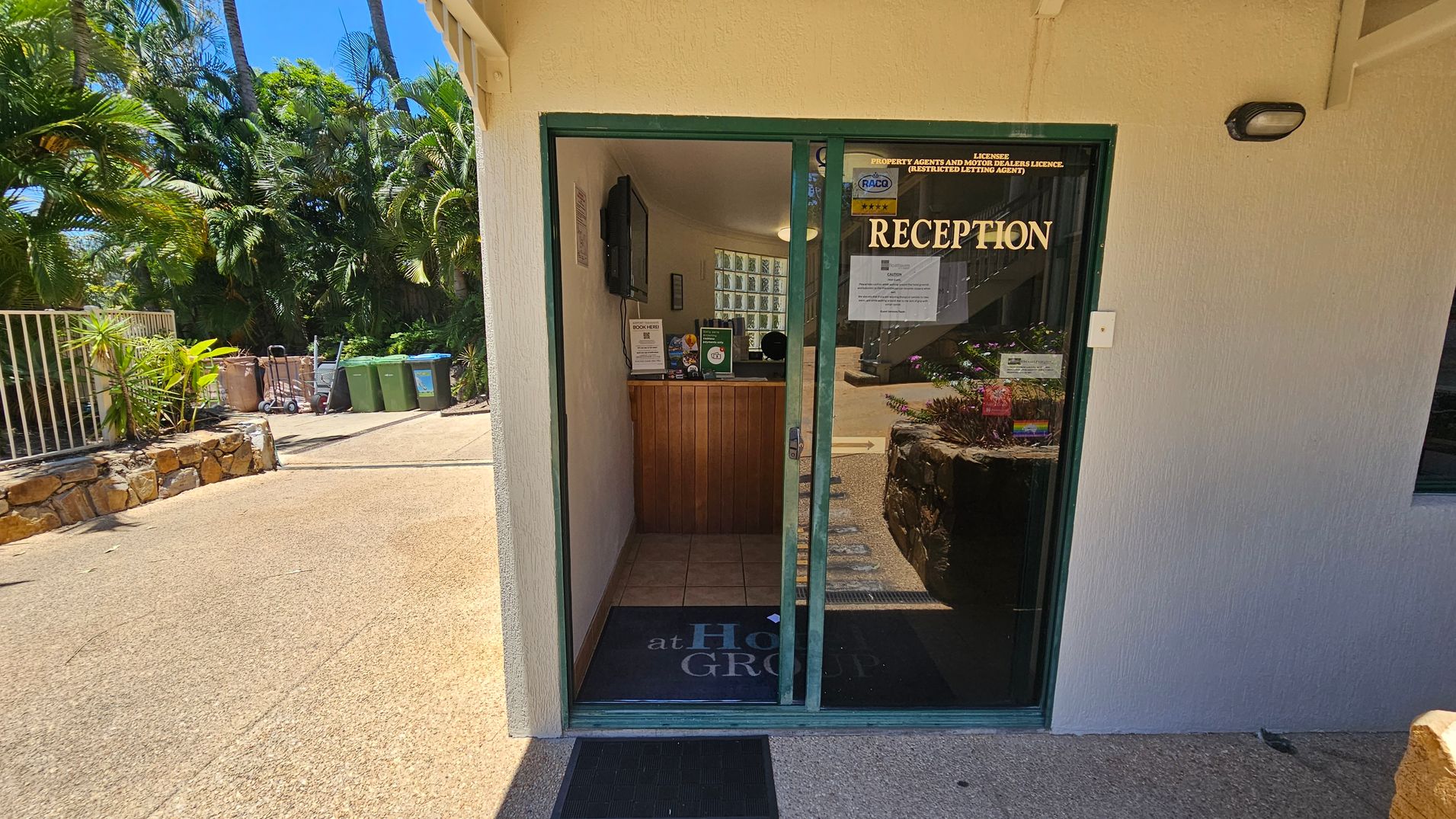 A small reception area with a glass door labelled "RECEPTION" in gold letters. Inside, there's a wooden counter with brochures and a computer screen. The floor is tiled. Outside, to the left, there are green and brown bins, a trolley, and lush greenery with palm trees. A stone wall lines the path leading to the entrance. The building exterior is light-coloured with a textured finish. A small reception area with a glass door labelled "RECEPTION" in gold letters. Inside, there's a wooden counter with brochures and a computer screen. The floor is tiled. Outside, to the left, there are green and brown bins, a trolley, and lush greenery with palm trees. A stone wall lines the path leading to the entrance. The building exterior is light-coloured with a textured finish.