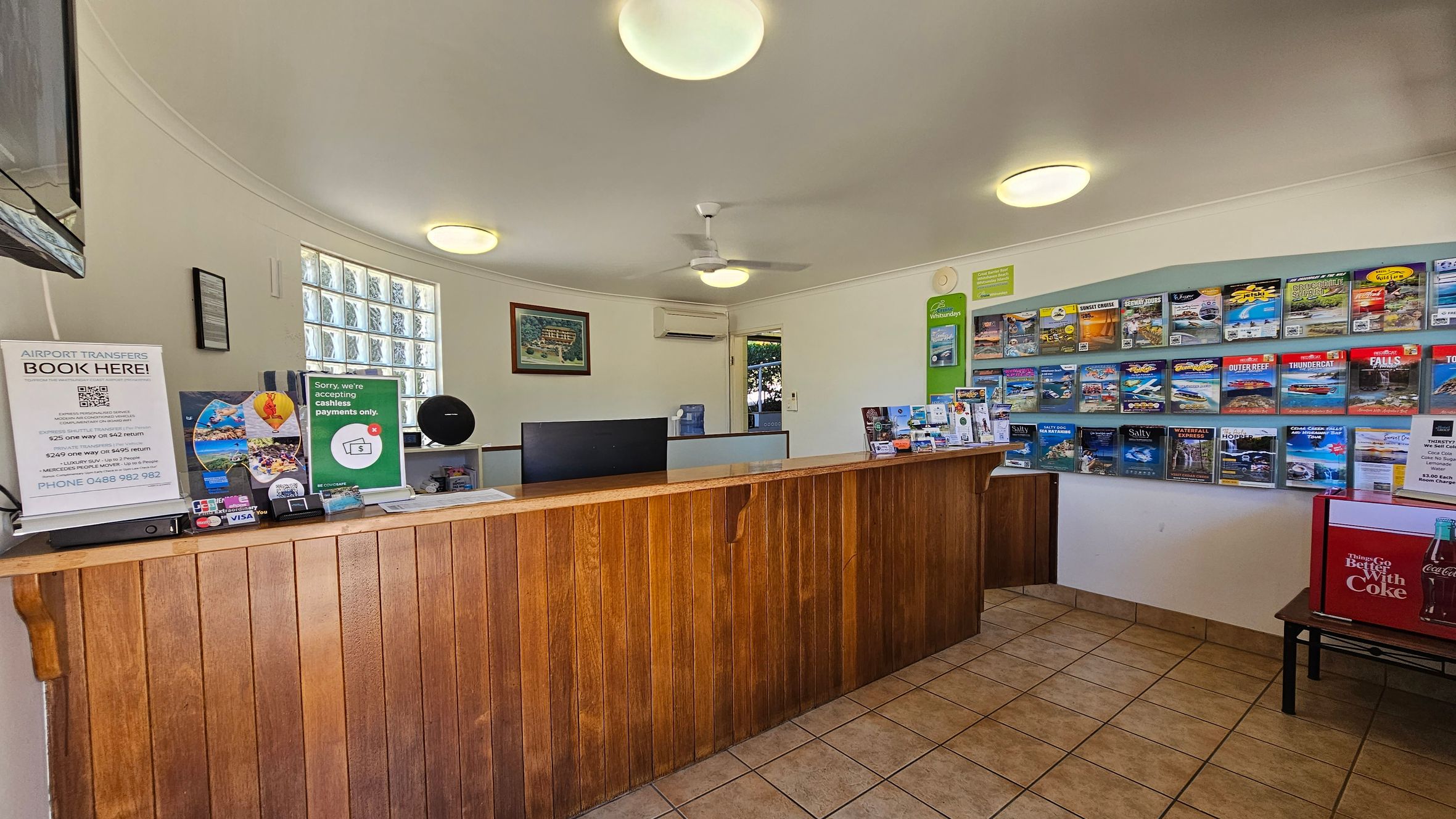 A reception area with a wooden counter in the foreground. On the left, there are signs for airport transfers and cashless payments. Behind the counter, a computer monitor and various brochures are visible. The right wall features a display of colourful brochures. The floor is tiled, and the ceiling has several round lights and a ceiling fan. A small Coca-Cola fridge is on the right side. A window with glass blocks is on the left wall, allowing natural light in. A reception area with a wooden counter in the foreground. On the left, there are signs for airport transfers and cashless payments. Behind the counter, a computer monitor and various brochures are visible. The right wall features a display of colourful brochures. The floor is tiled, and the ceiling has several round lights and a ceiling fan. A small Coca-Cola fridge is on the right side. A window with glass blocks is on the left wall, allowing natural light in.