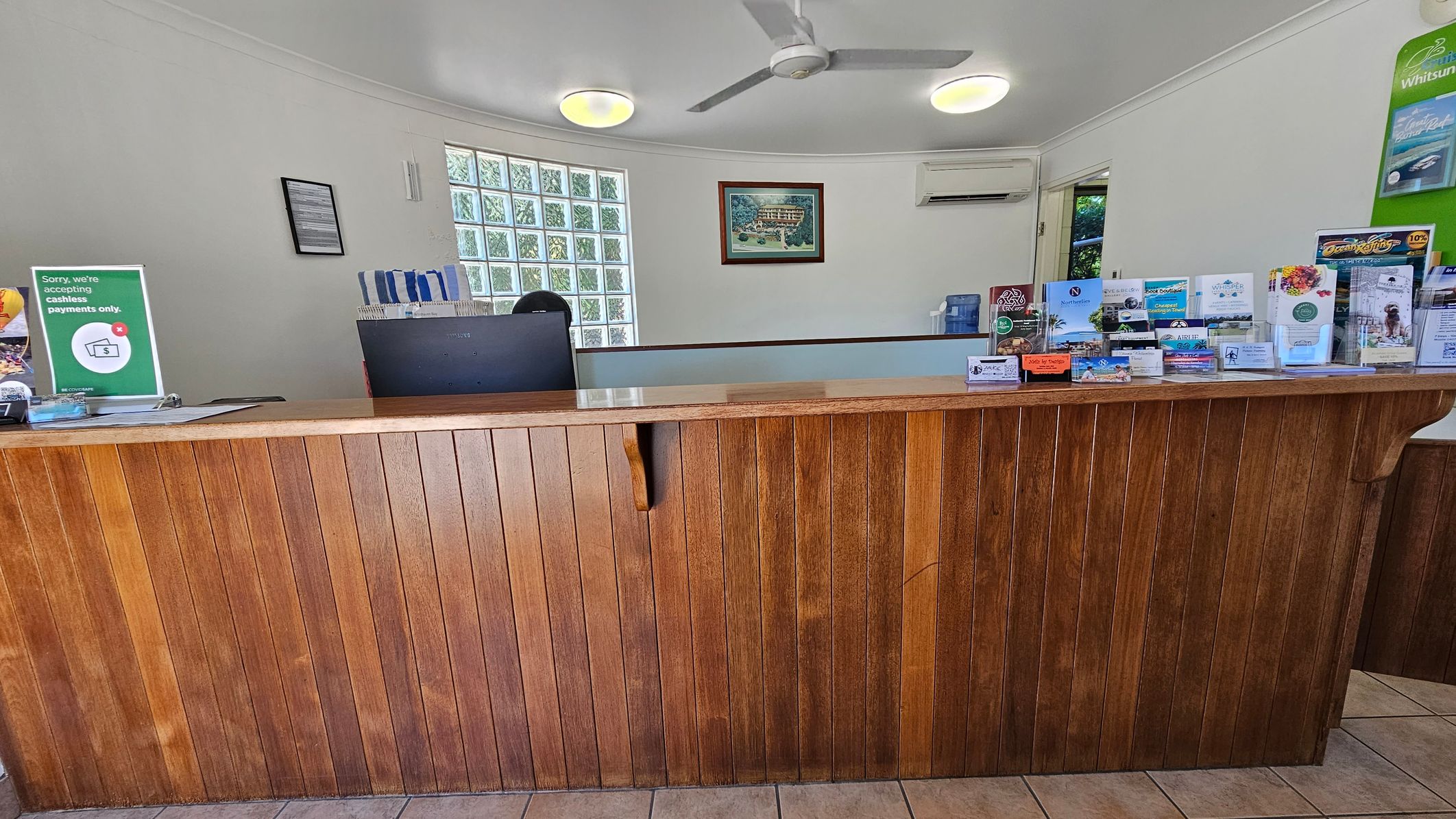 A reception area with a wooden counter in the foreground. On the right side of the counter, there are various brochures and business cards displayed. Behind the counter, a computer monitor is visible. The background features a wall with a framed picture, an air conditioning unit, and a glass block window. The ceiling has two round lights and a ceiling fan. The floor is tiled. A reception area with a wooden counter in the foreground. On the right side of the counter, there are various brochures and business cards displayed. Behind the counter, a computer monitor is visible. The background features a wall with a framed picture, an air conditioning unit, and a glass block window. The ceiling has two round lights and a ceiling fan. The floor is tiled.