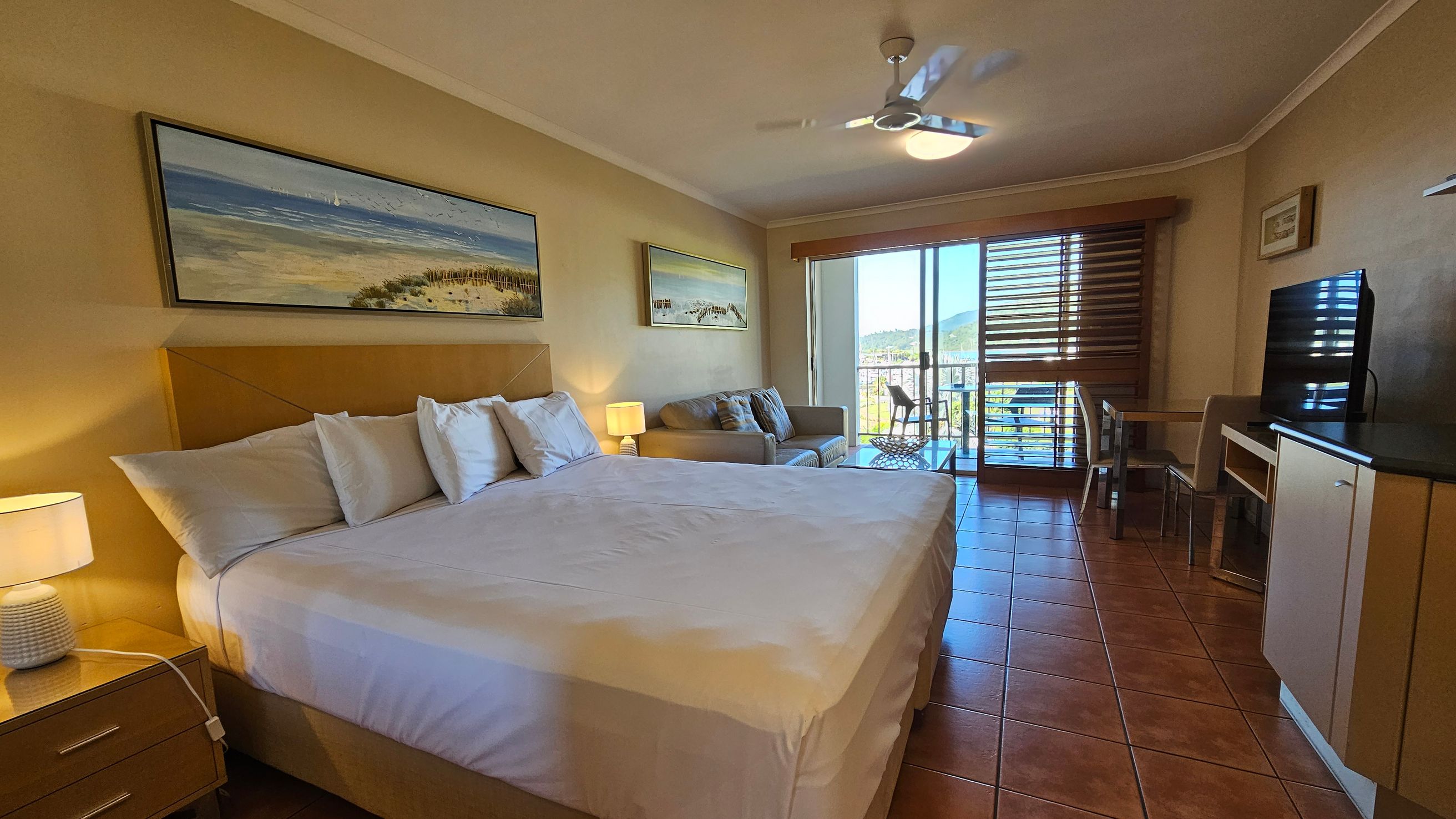 A hotel room with a large bed on the left, adorned with white pillows and sheets. A bedside table with a lamp is next to the bed. Above the bed, a landscape painting hangs on the wall. In the middle, there's a beige sofa with cushions and another painting above it. On the right, a flat-screen TV sits on a cabinet, with a small table and chair nearby. The room has a tiled floor and a ceiling fan. Sliding glass doors lead to a balcony with a view of hills and water. A hotel room with a large bed on the left, adorned with white pillows and sheets. A bedside table with a lamp is next to the bed. Above the bed, a landscape painting hangs on the wall. In the middle, there's a beige sofa with cushions and another painting above it. On the right, a flat-screen TV sits on a cabinet, with a small table and chair nearby. The room has a tiled floor and a ceiling fan. Sliding glass doors lead to a balcony with a view of hills and water.