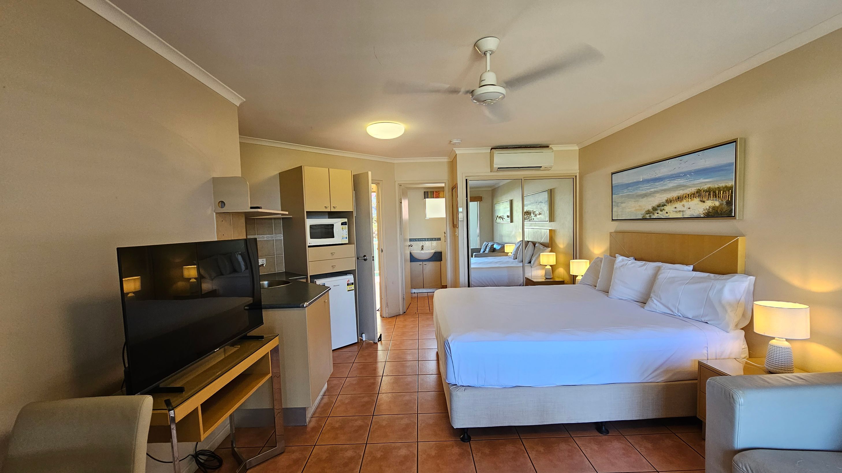 A hotel room with terracotta-tiled flooring and cream walls. On the right, there's a large bed with white linens, flanked by bedside tables with lamps. Above the bed is a framed beach painting. On the left, a flat-screen TV sits on a wooden stand. A kitchenette with a microwave, fridge, and sink is visible. In the background, a doorway leads to a bathroom. A ceiling fan and air conditioning unit are mounted above. A hotel room with terracotta-tiled flooring and cream walls. On the right, there's a large bed with white linens, flanked by bedside tables with lamps. Above the bed is a framed beach painting. On the left, a flat-screen TV sits on a wooden stand. A kitchenette with a microwave, fridge, and sink is visible. In the background, a doorway leads to a bathroom. A ceiling fan and air conditioning unit are mounted above.