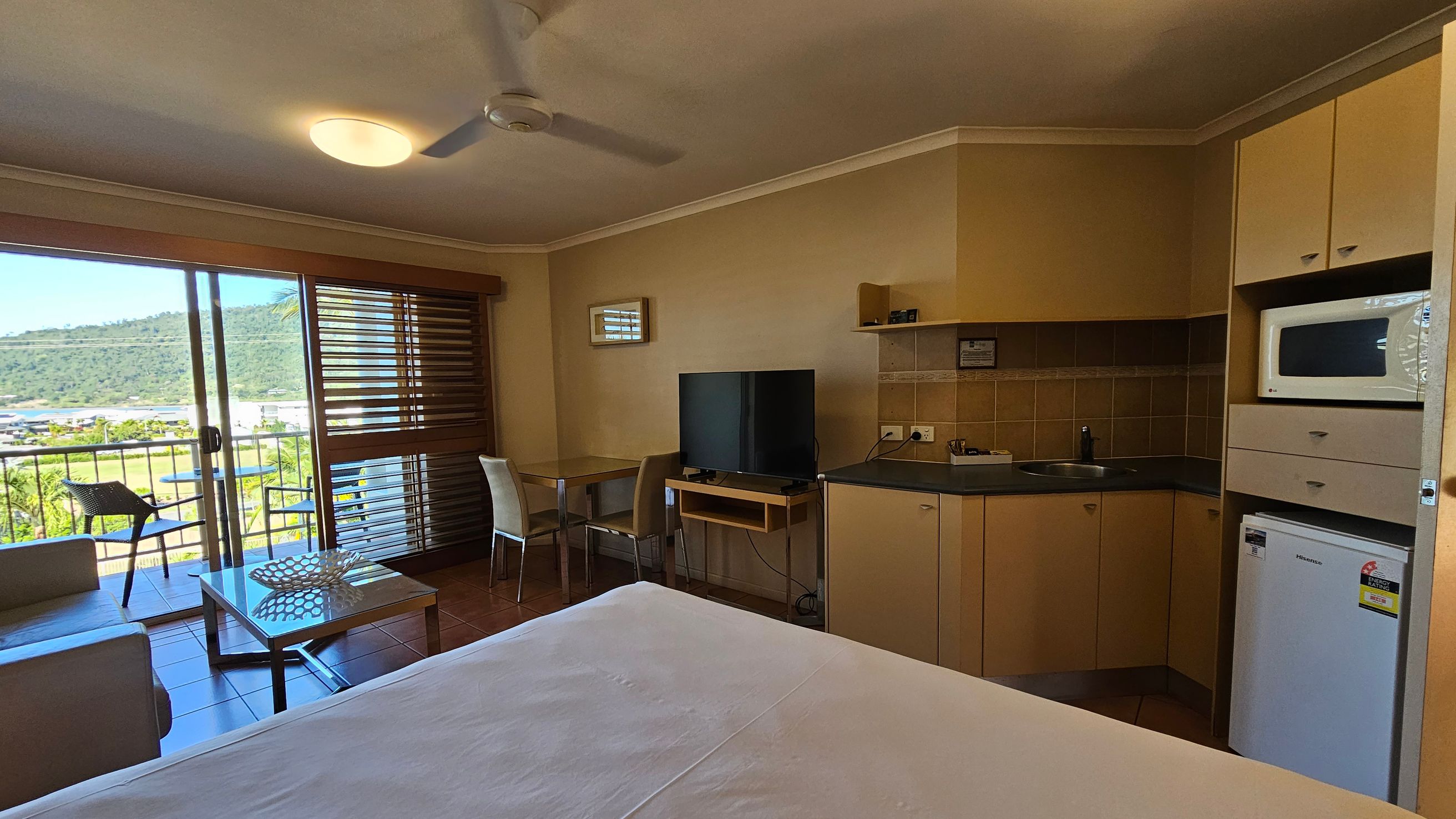 A room with a view of a balcony and hills in the background. On the left, there's a sliding glass door with wooden blinds, leading to a small balcony with a chair. Inside, a sofa and a coffee table are near the door. In the middle, a small dining table with two chairs is next to a flat-screen TV on a stand. On the right, a kitchenette with a microwave, sink, and mini-fridge. The ceiling has a fan and a light fixture, and the floor is tiled. A room with a view of a balcony and hills in the background. On the left, there's a sliding glass door with wooden blinds, leading to a small balcony with a chair. Inside, a sofa and a coffee table are near the door. In the middle, a small dining table with two chairs is next to a flat-screen TV on a stand. On the right, a kitchenette with a microwave, sink, and mini-fridge. The ceiling has a fan and a light fixture, and the floor is tiled.